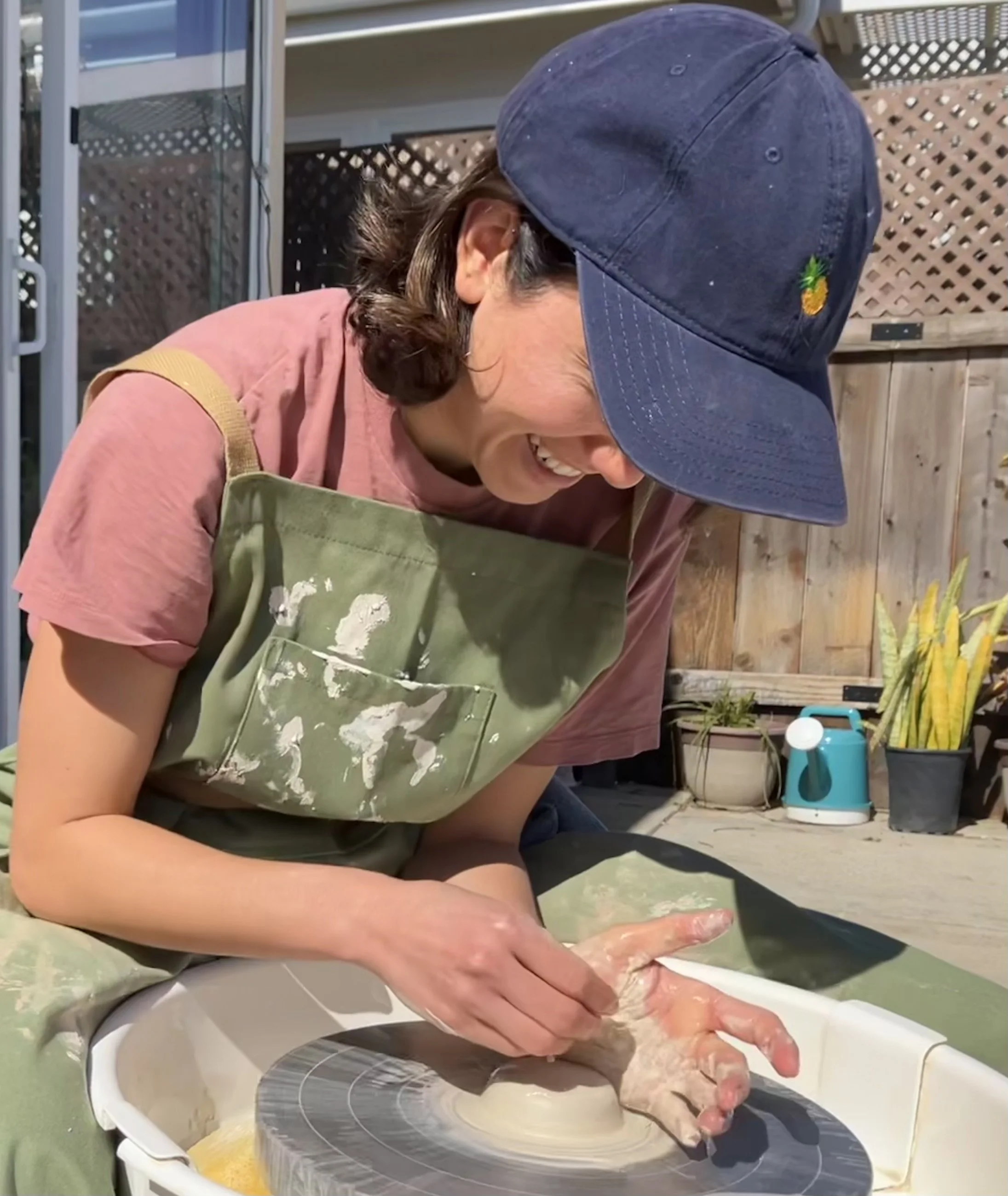A woman laughing while shaping clay on a pottery wheel outdoors, wearing a blue cap, a pink t-shirt, and a green apron splattered with white paint.