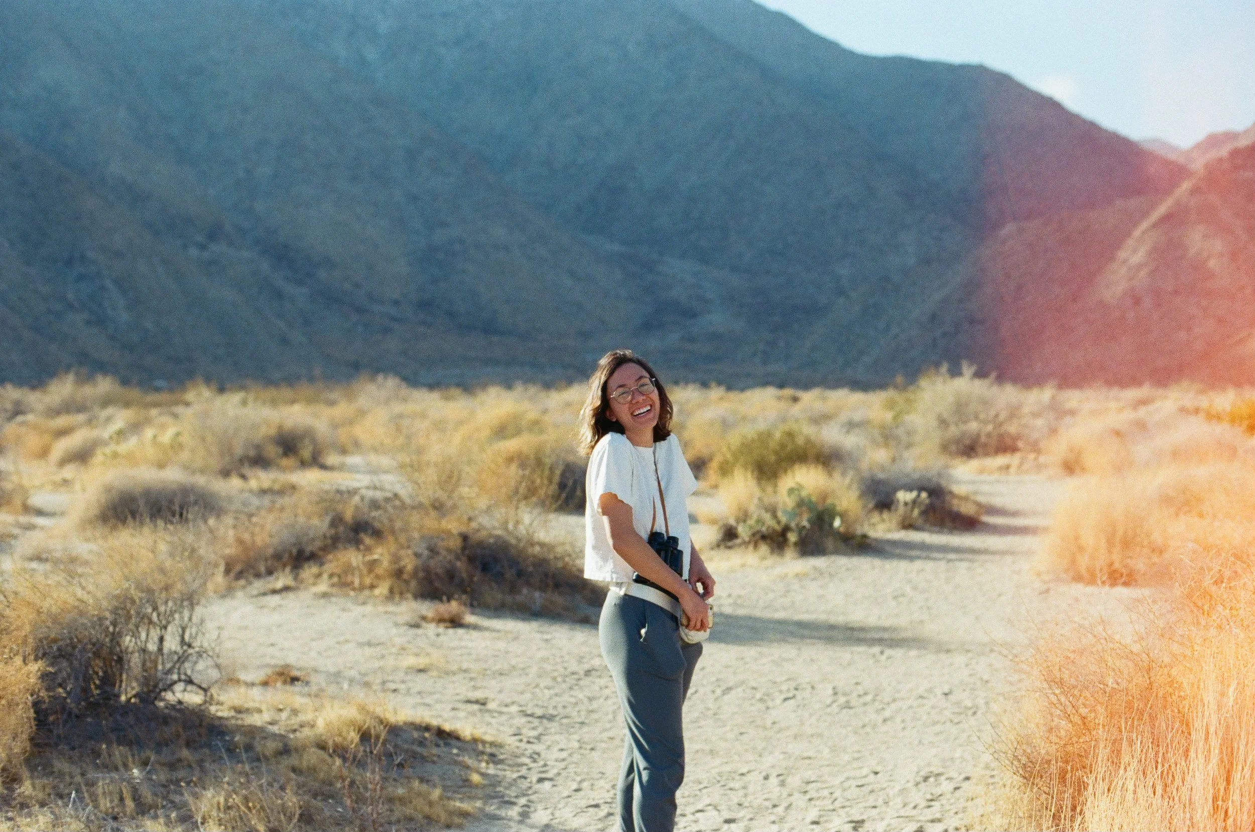 A woman with glasses standing and smiling in a desert landscape with mountains in the background