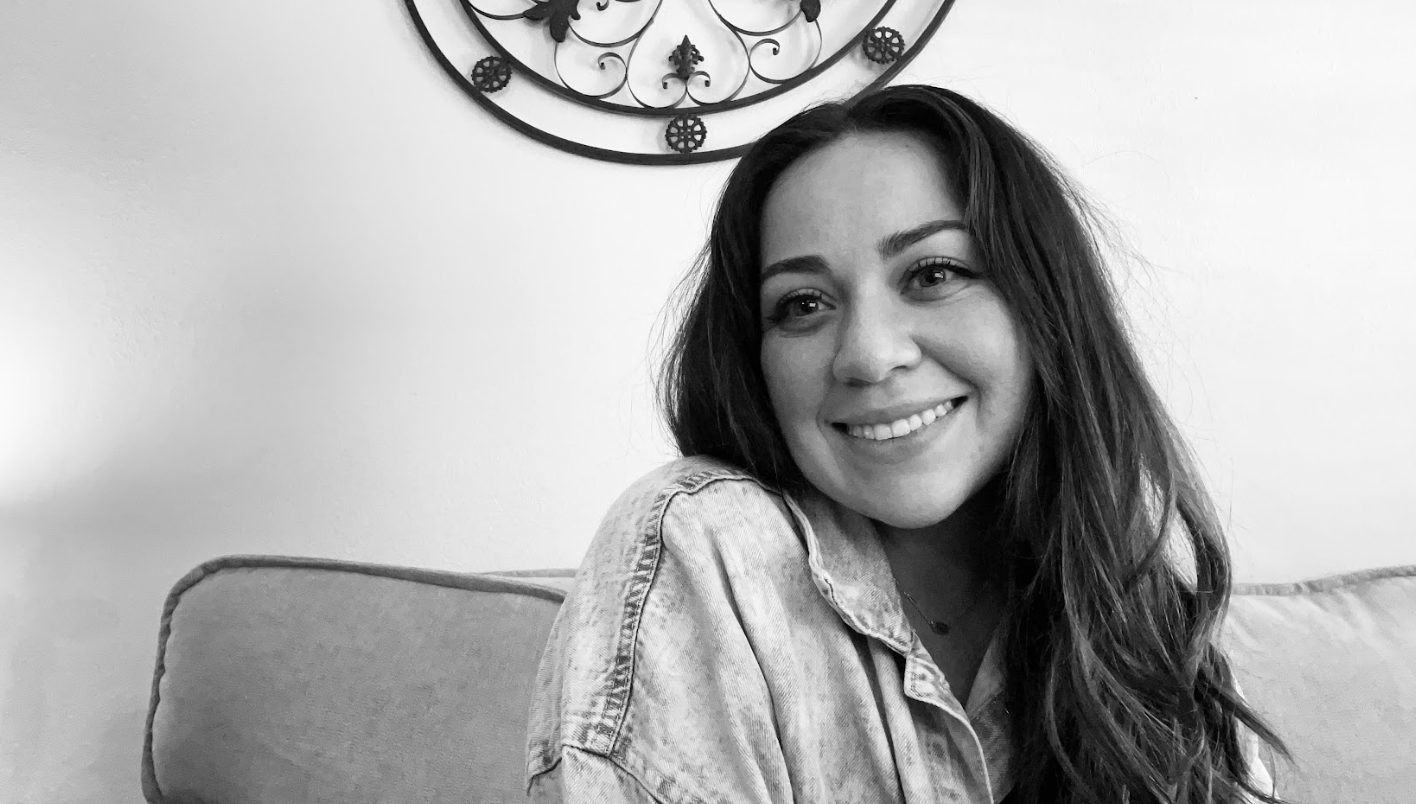 Black and white photo of a smiling woman with long, wavy hair, sitting on a sofa with a decorative wall art piece in the background.