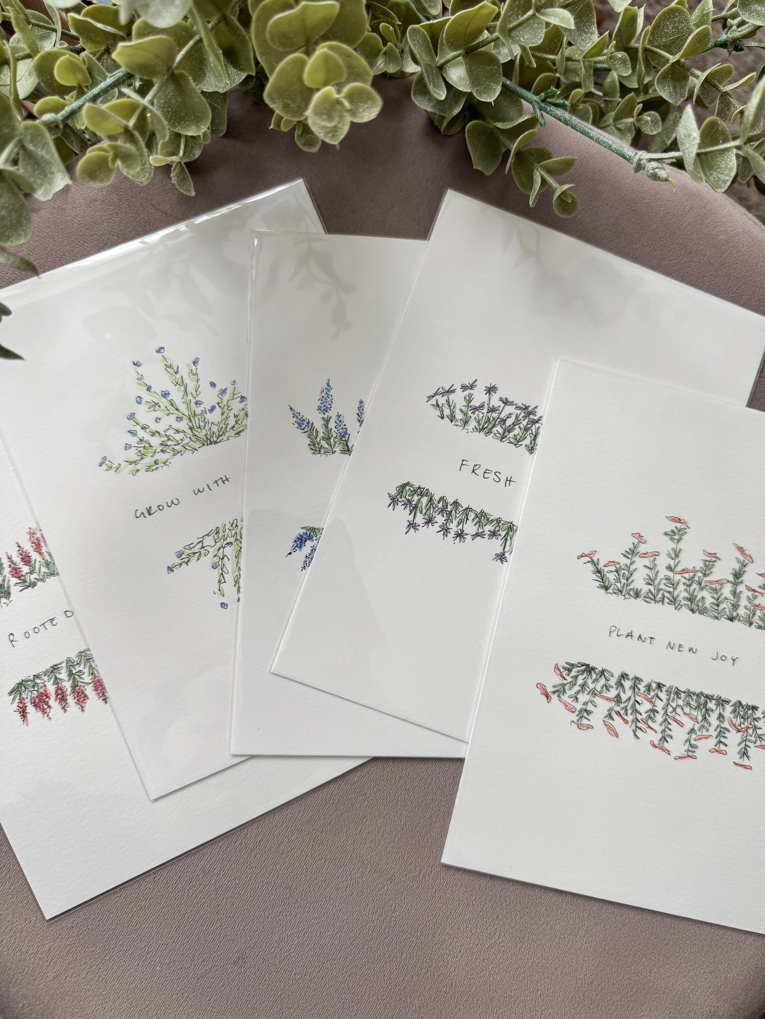Four white cards with colorful floral illustrations and inspirational phrases, arranged over a pinkish surface with green foliage in the background.