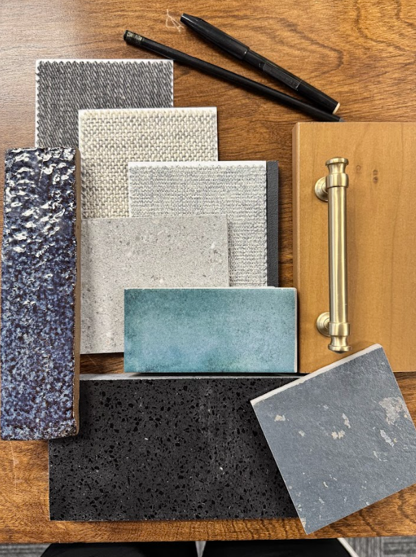 Collection of various textured tile samples in shades of gray, blue, beige, and black, along with a black pen and a brass door handle on a wooden surface.