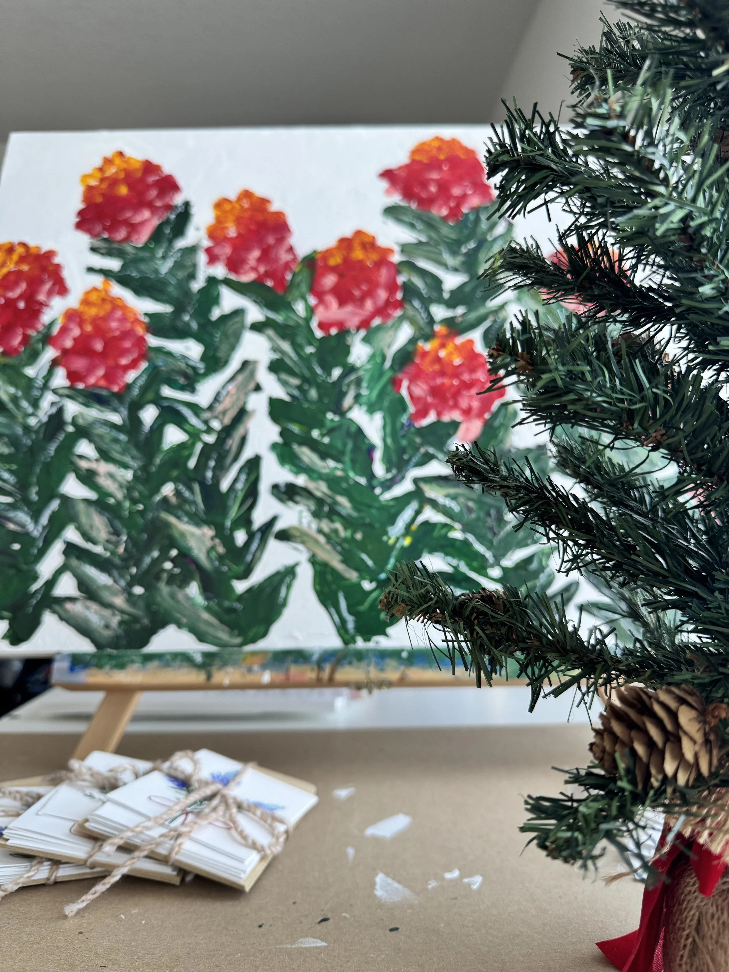 A colorful painting of red and orange flowers with green leaves on an easel, with a partially visible Christmas tree with pine cones and red ribbon in the foreground, and some wrapped cards on a table.