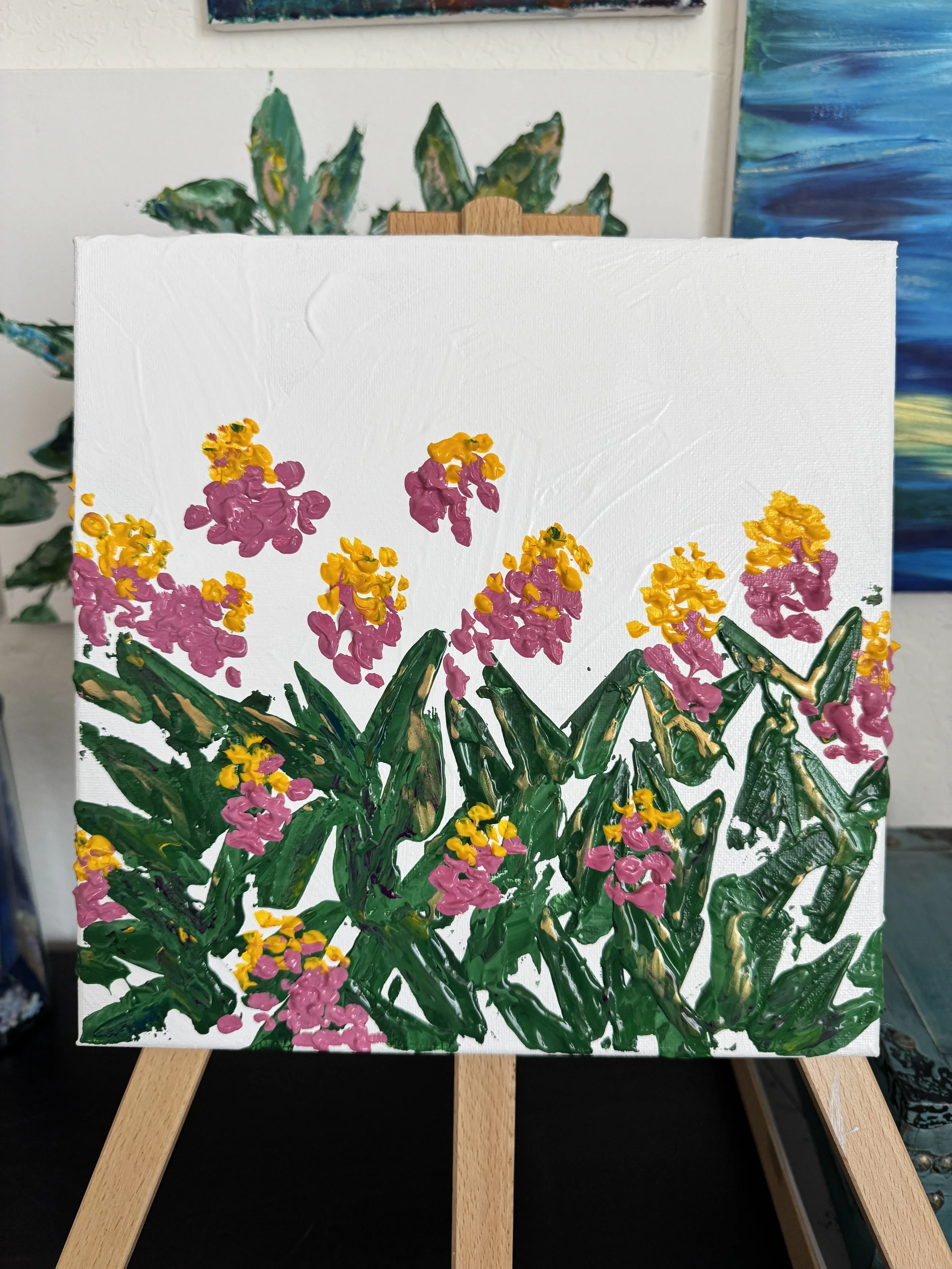 A small canvas painting of a vibrant floral scene, with pink and yellow flowers and green leaves, displayed on a wooden easel.
