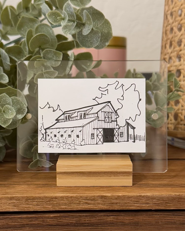 A black and white line drawing of a barn with a silo, trees, and a fence, held inside a clear acrylic frame with a wooden base. Background includes green plants and a pink container.