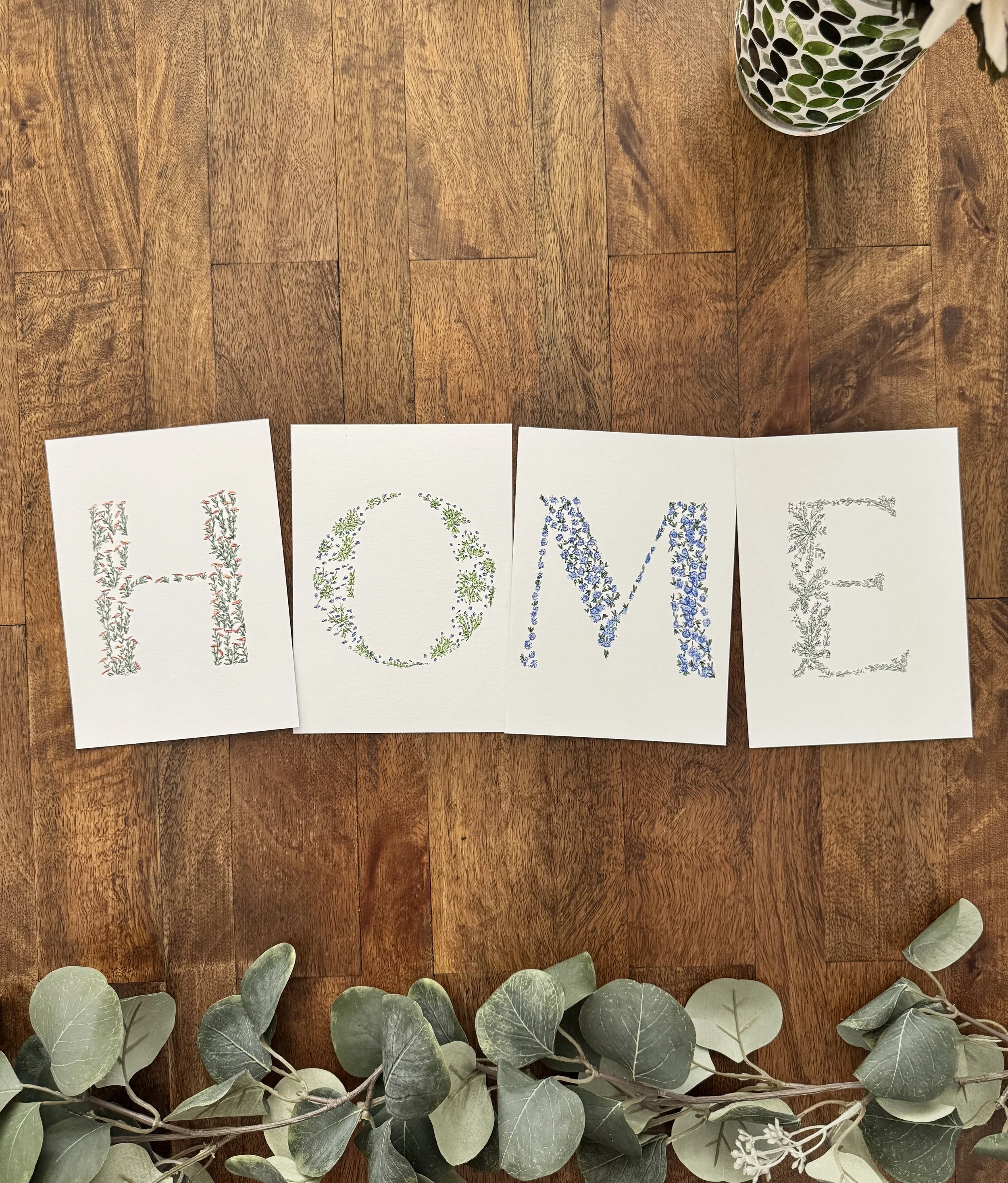 Four cards arranged on a wooden floor spell out the word 'HOME' with decorative floral letters. A small plant with green leaves is on the top right corner, and eucalyptus leaves are at the bottom of the image.
