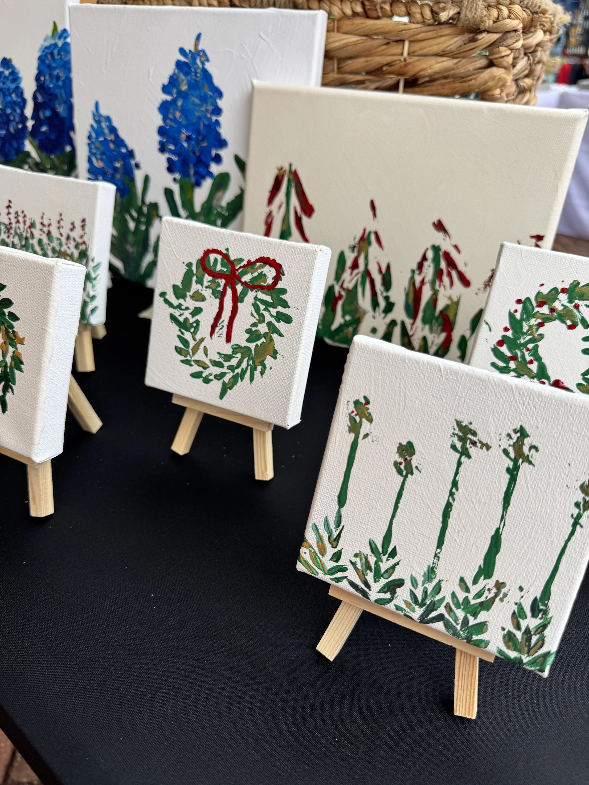 Small canvas paintings of Christmas-themed wreaths and holiday plants on miniature easels, displayed on a black table.