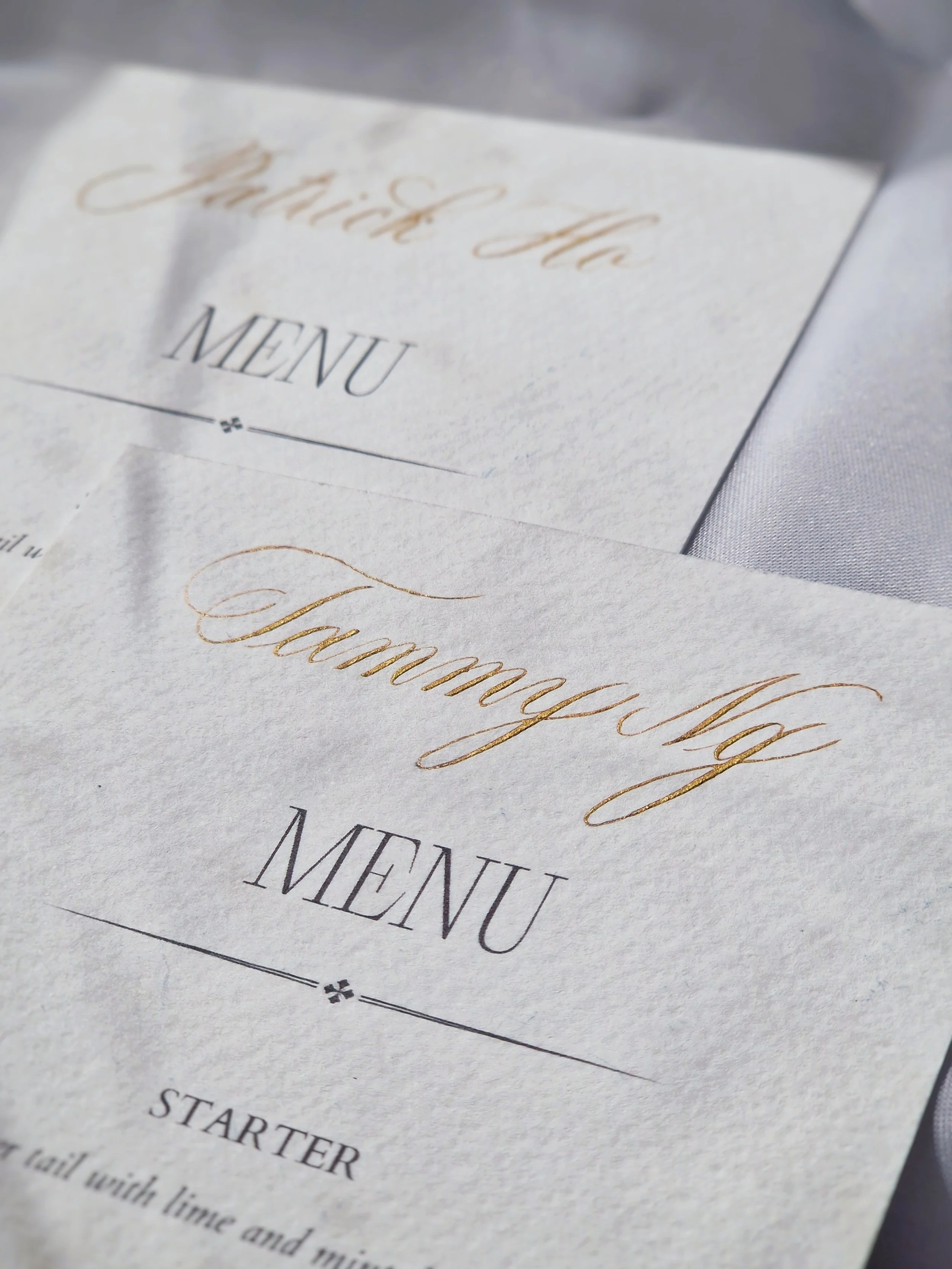 Close-up of a wedding menu card with elegant gold cursive text and black serif text, placed on a white tablecloth.