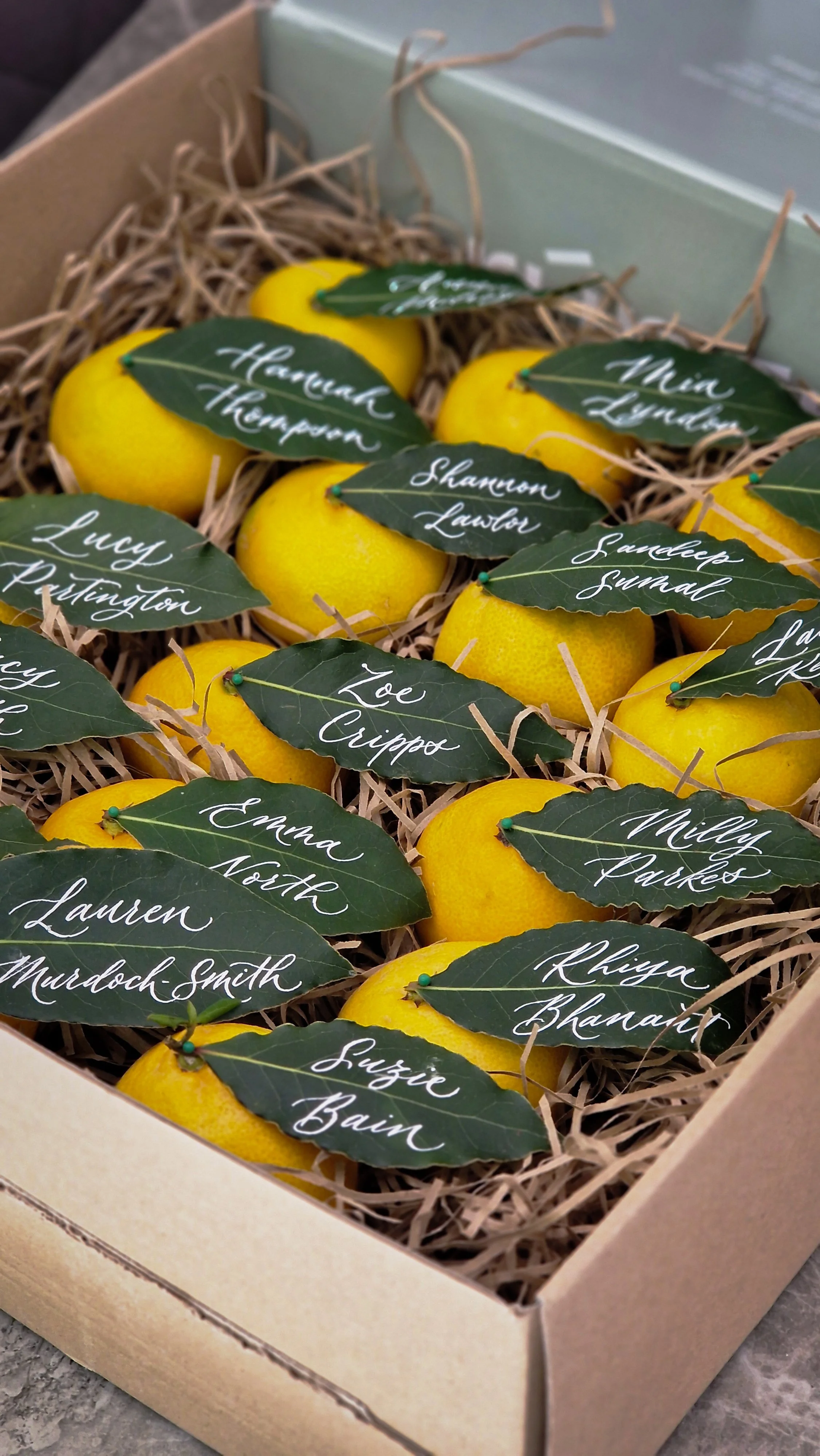 A box of yellow lemons decorated with green leaves that have handwritten white names on them.