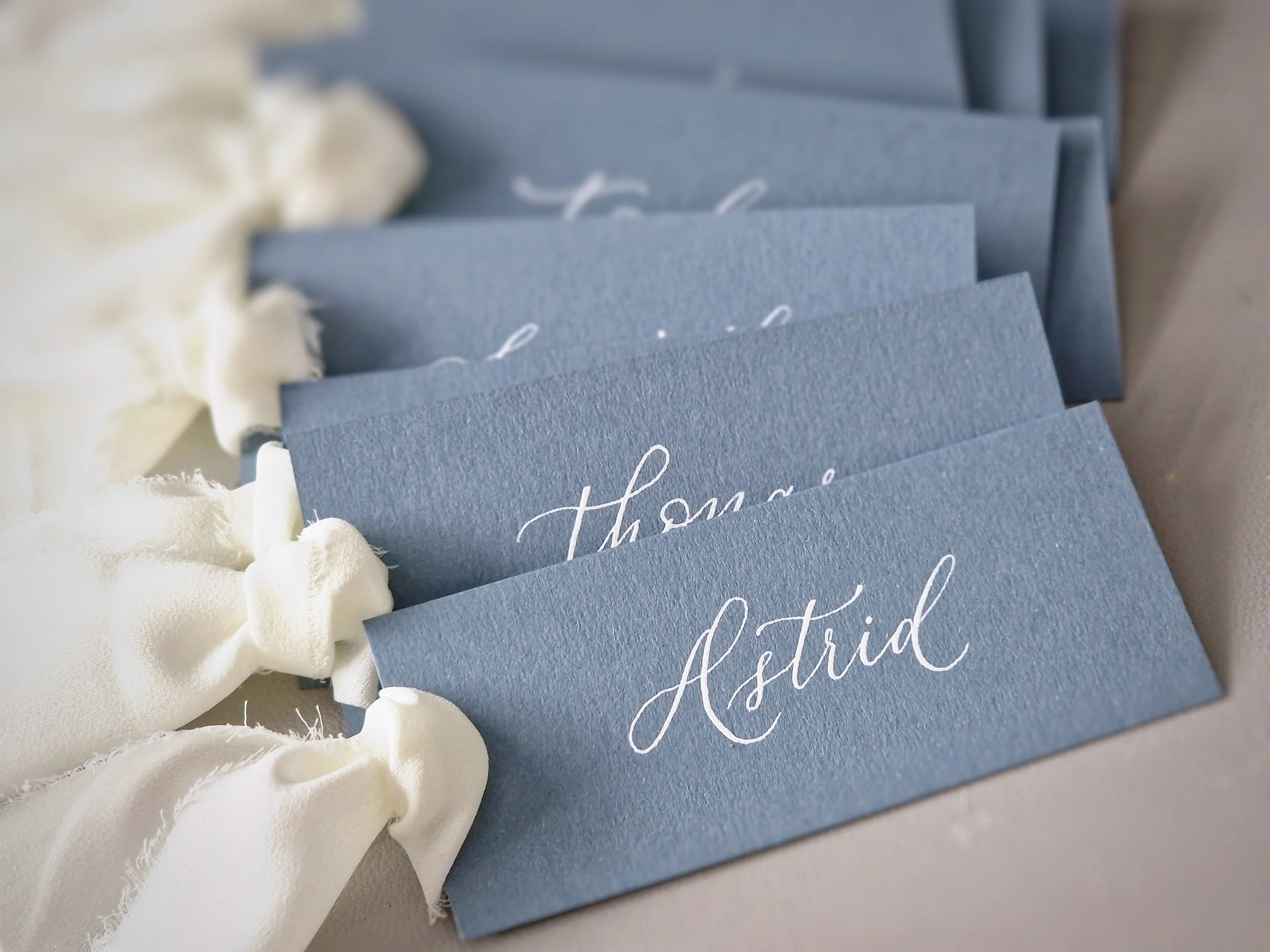 A group of blue place cards with the name 'Astrid' written in white cursive ink, arranged in a row on a table, with white fabric or ribbon decorations on the side.