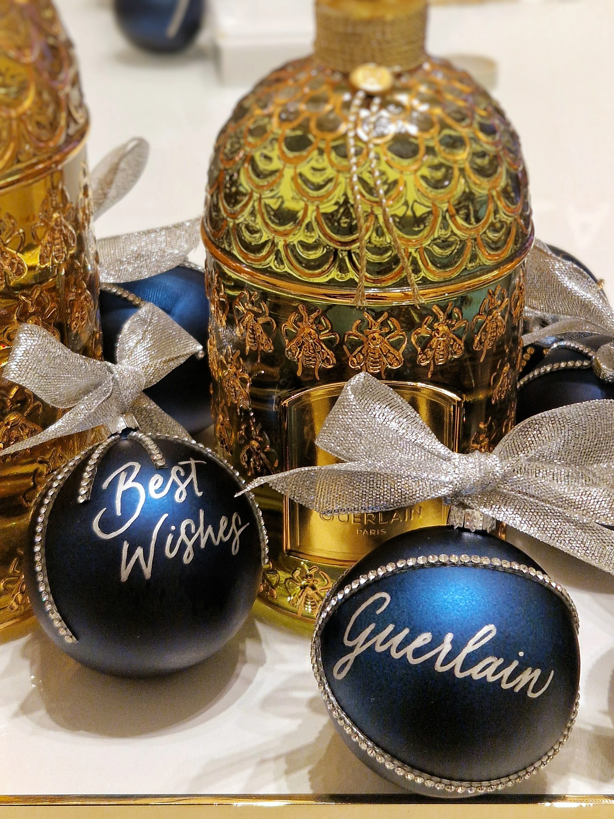Silver calligraphy on blue Christmas baubles decorated with silver rhinestones and ribbon. There is a gold honeycomb and bee Guerlain perfume bottle in the background