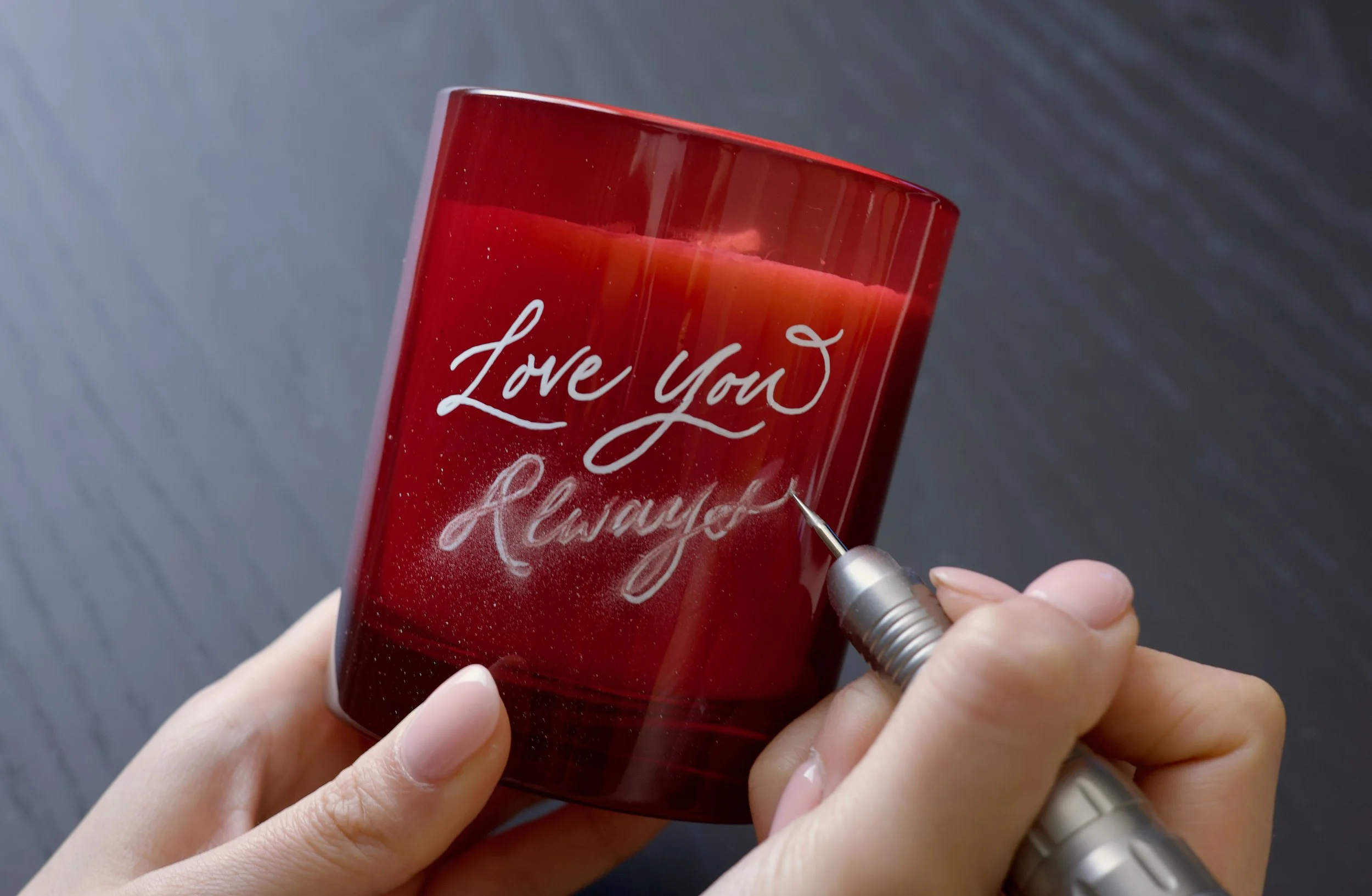A person engraving on a red glass candle holder with the words 'Love You Always'.