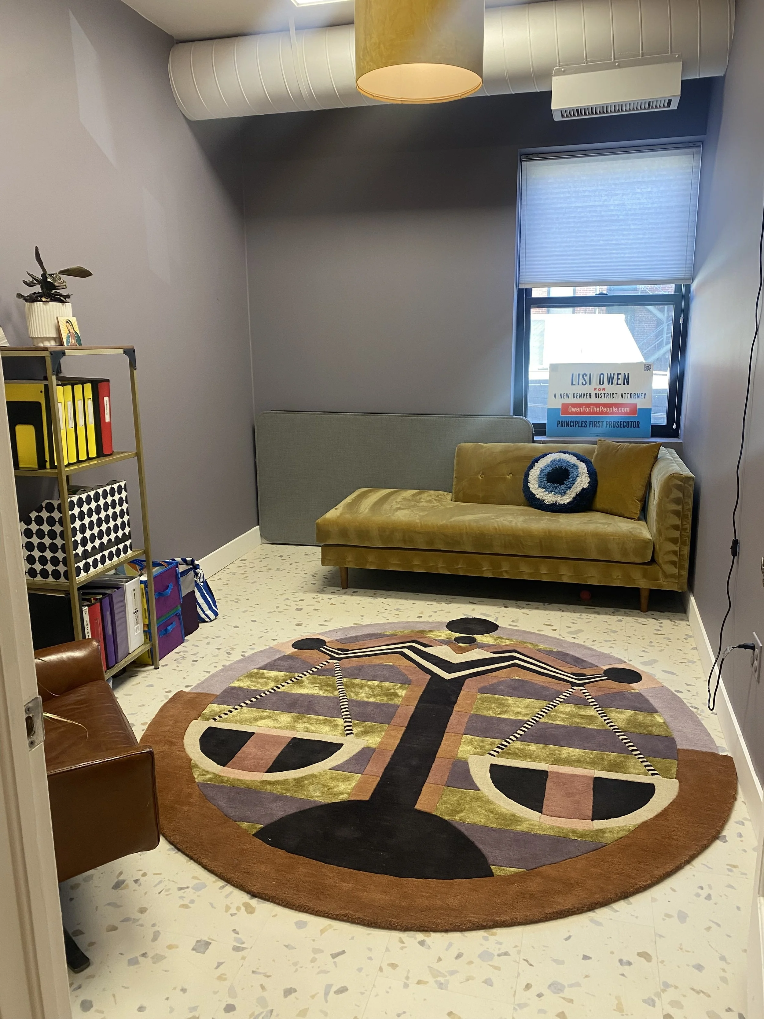 A small, modern waiting room or office space with a circular colorful rug on the floor, a mustard yellow sofa with a decorative pillow, a brown armchair, a tall shelving unit with binders and decor, and a window with an election sign for Lisi Owen, candidate for a Denver district attorney, on the window.