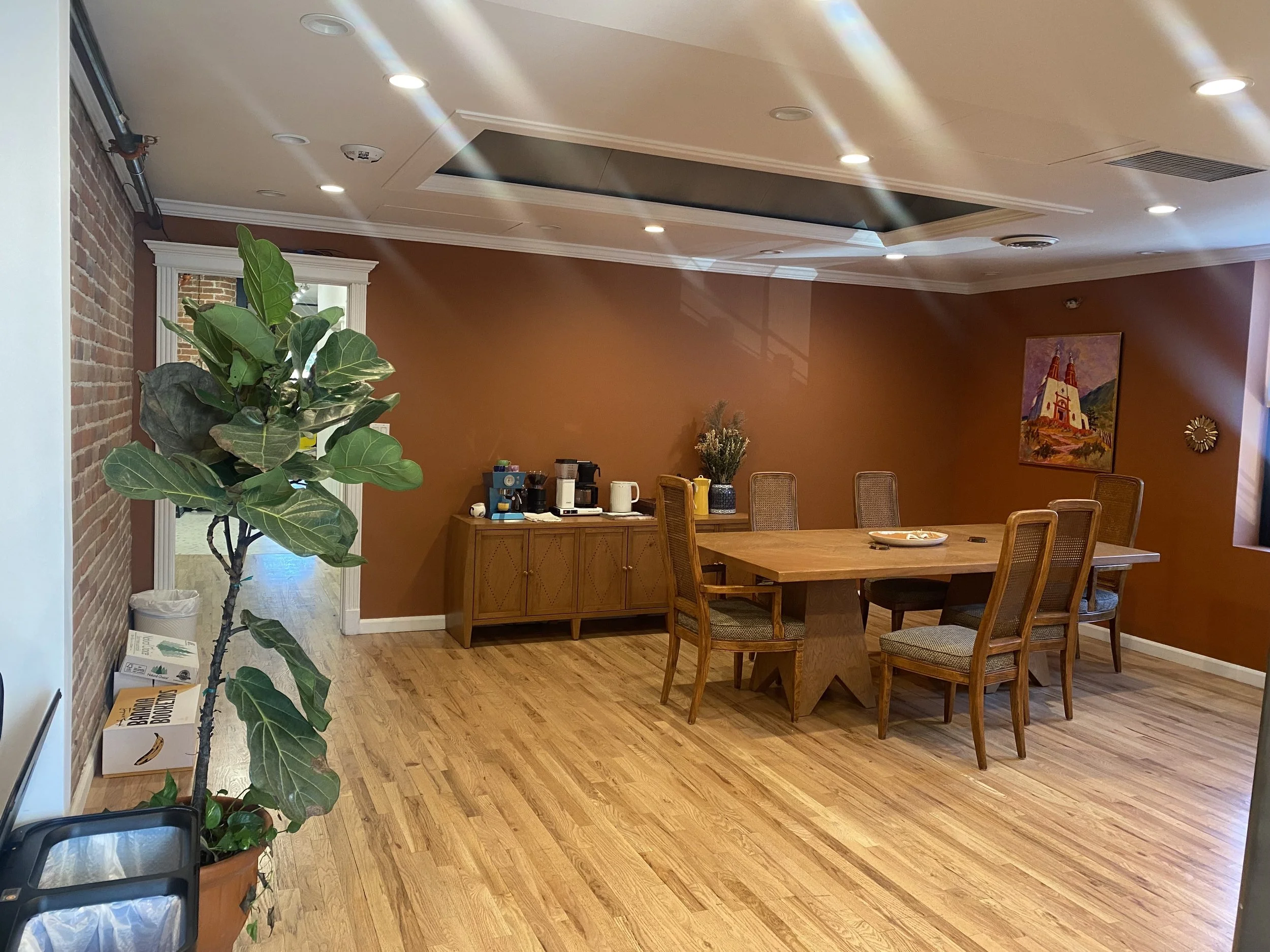 Dining room with a wooden table and six chairs, a sideboard with coffee maker and supplies, a large potted plant, and artwork on the walls. The room has hardwood floors and a brick wall on the left, with sunlight coming through a window.