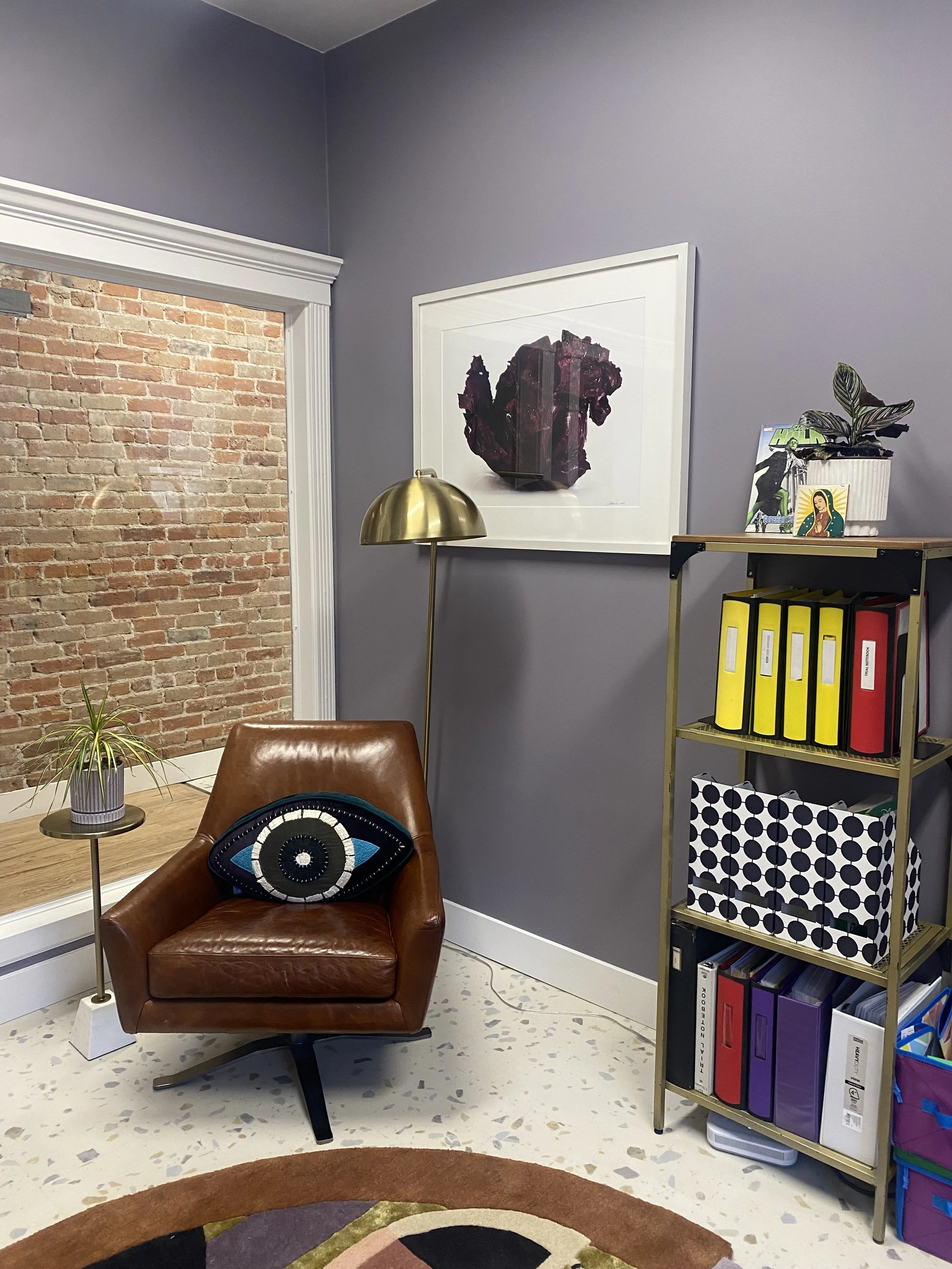Interior of a cozy office corner with a brown leather armchair, an eye-shaped cushion, a tall gold floor lamp, a small wooden side table with a potted plant, a purple wall, a framed abstract art piece, and a gold shelving unit with colorful binders and decorative items.