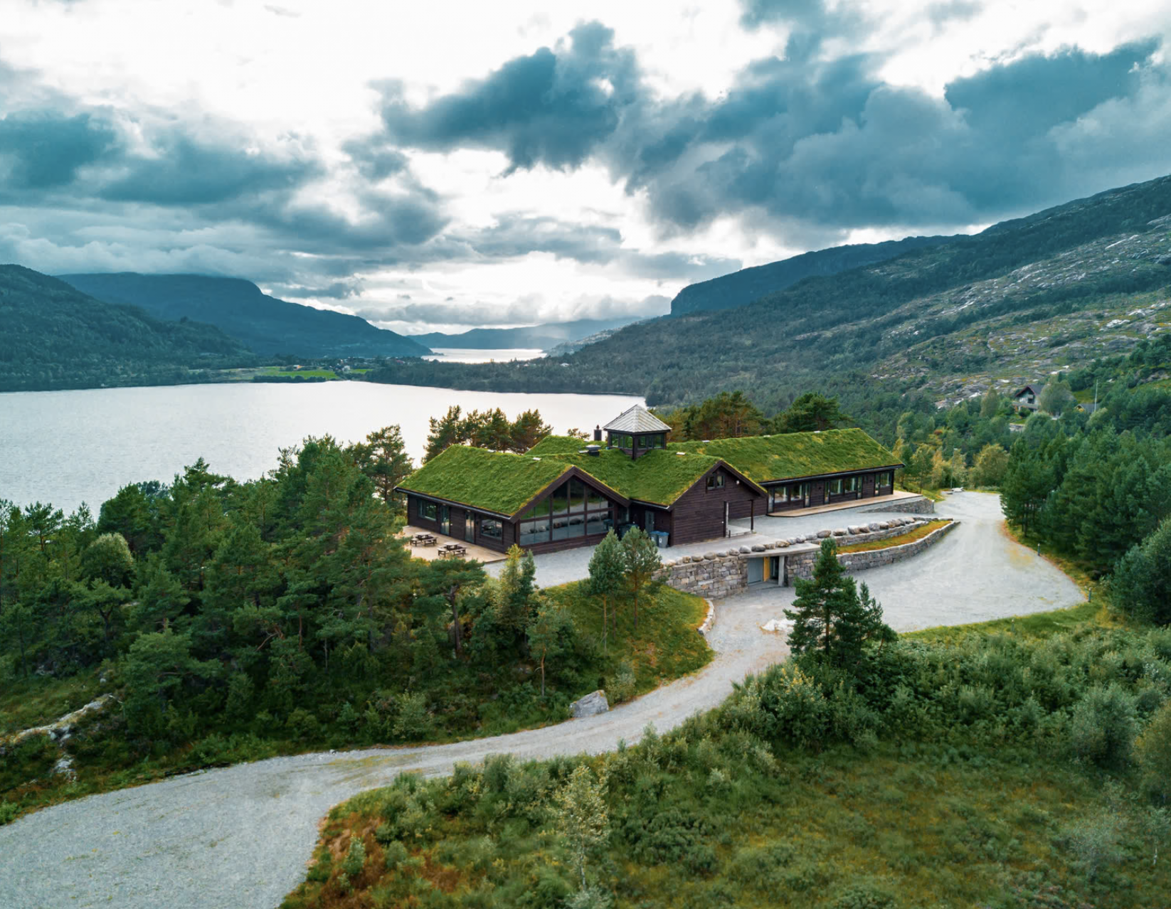 A modern house with a green roof situated among trees, overlooking a lake with mountains in the background during daytime with cloudy sky.