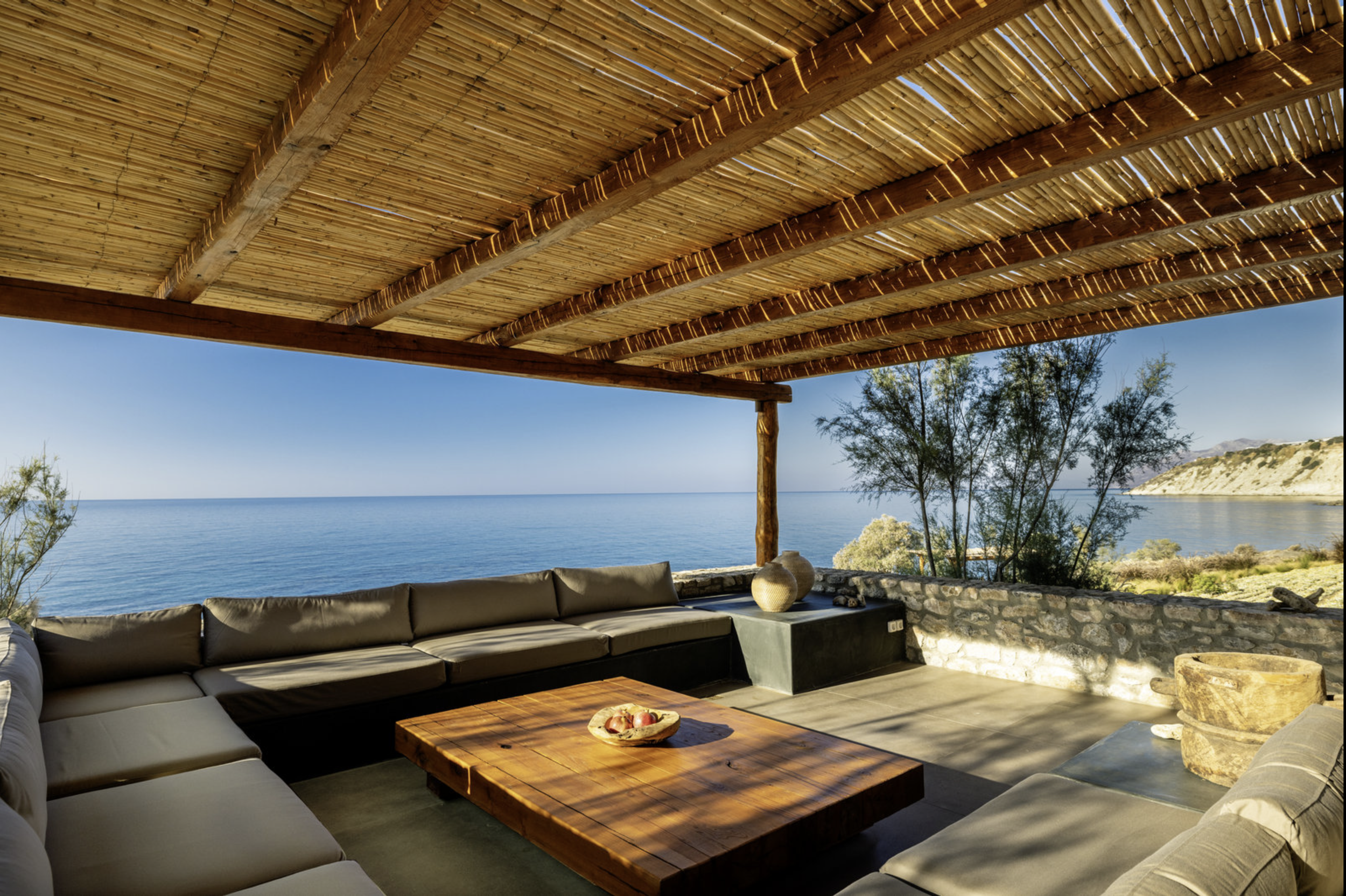 Outdoor seating area with sectional sofa and wooden table overlooking a calm sea, with trees and hills in the background, under a thatched roof canopy.