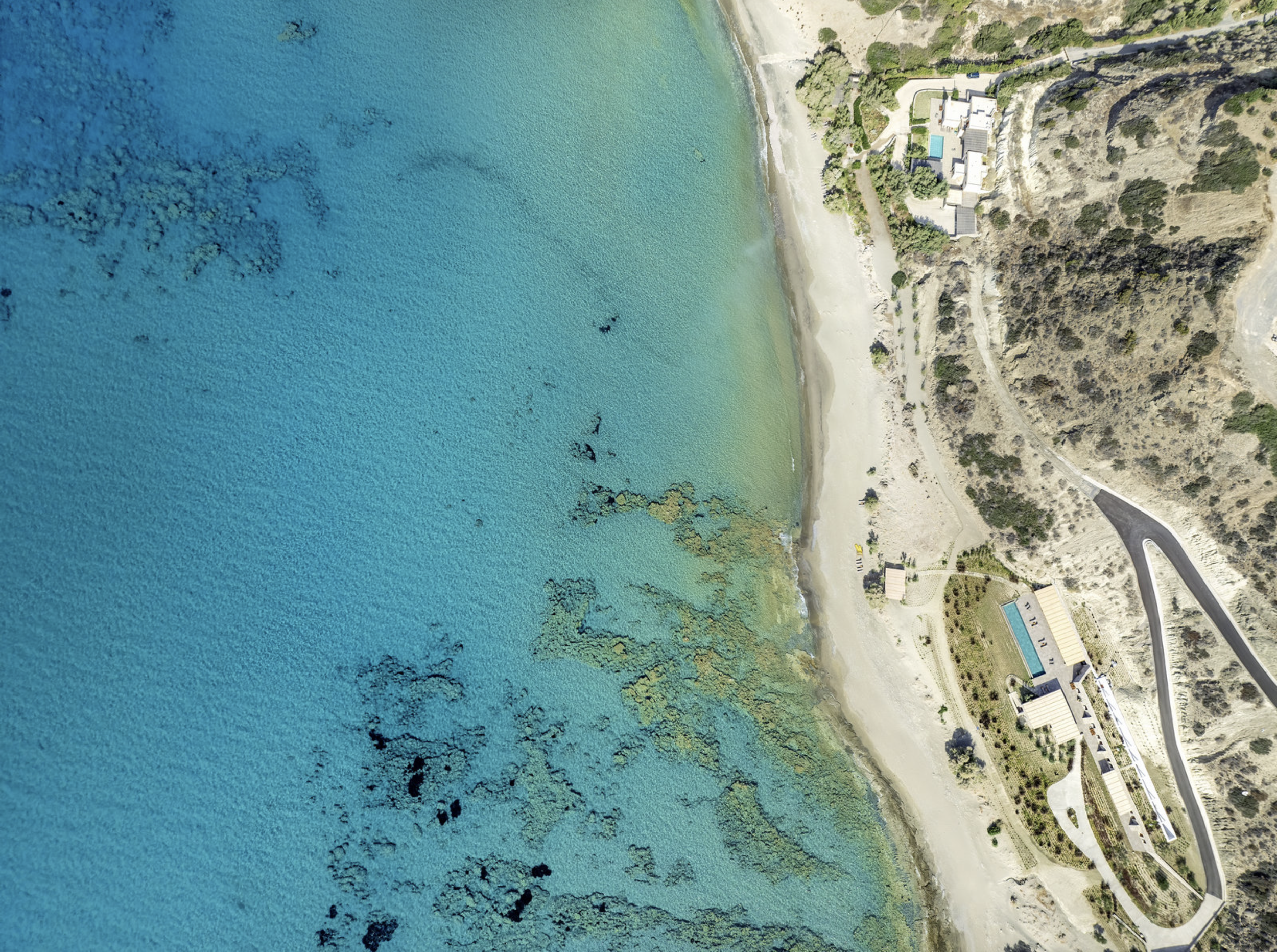 Overhead aerial view of a scenic coastal area featuring a beach with light sand, clear turquoise water with dark patches of underwater rocks or seaweed, and resort buildings with pools and roads on a hilly terrain.