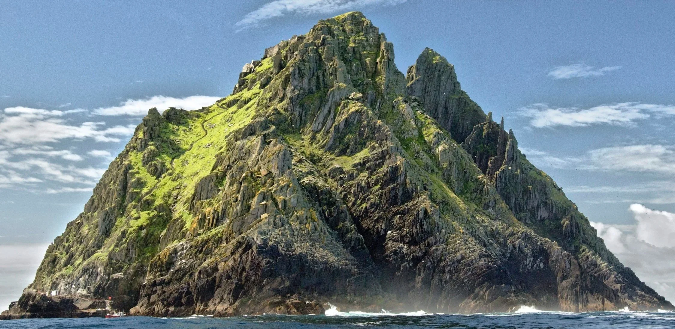 A large rocky island with steep, rugged cliffs covered in green vegetation. The island rises sharply from the ocean, with a partly cloudy sky in the background.