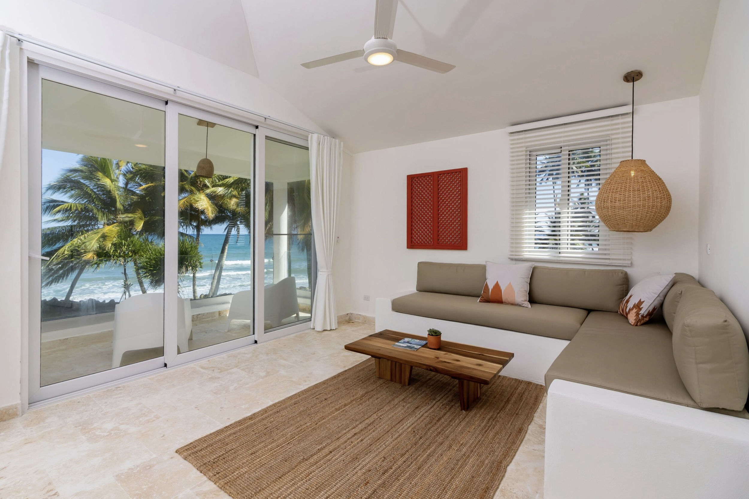 A living room with a large desert-colored L-shaped sofa, a small wooden coffee table, a window with white blinds, and a sliding glass door leading to a balcony overlooking a beach with palm trees and ocean waves.