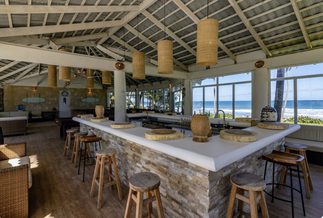 Interior of a beachside restaurant or bar with a white stone counter, wooden stools, wicker pendant lights, large windows showing the ocean, and palm trees outside.