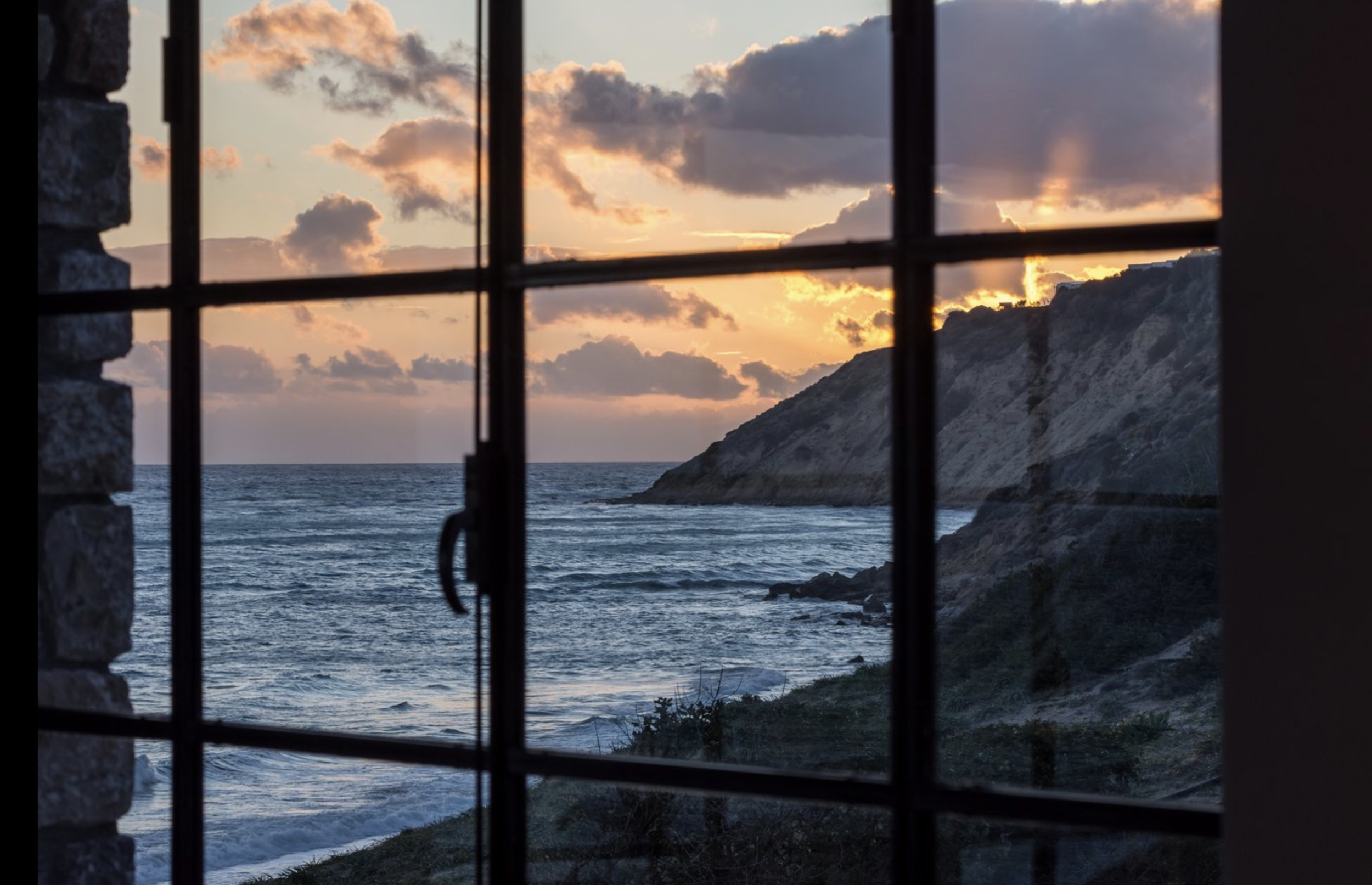 Sunset view of the ocean with clouds and a rocky coastline, seen through a window.