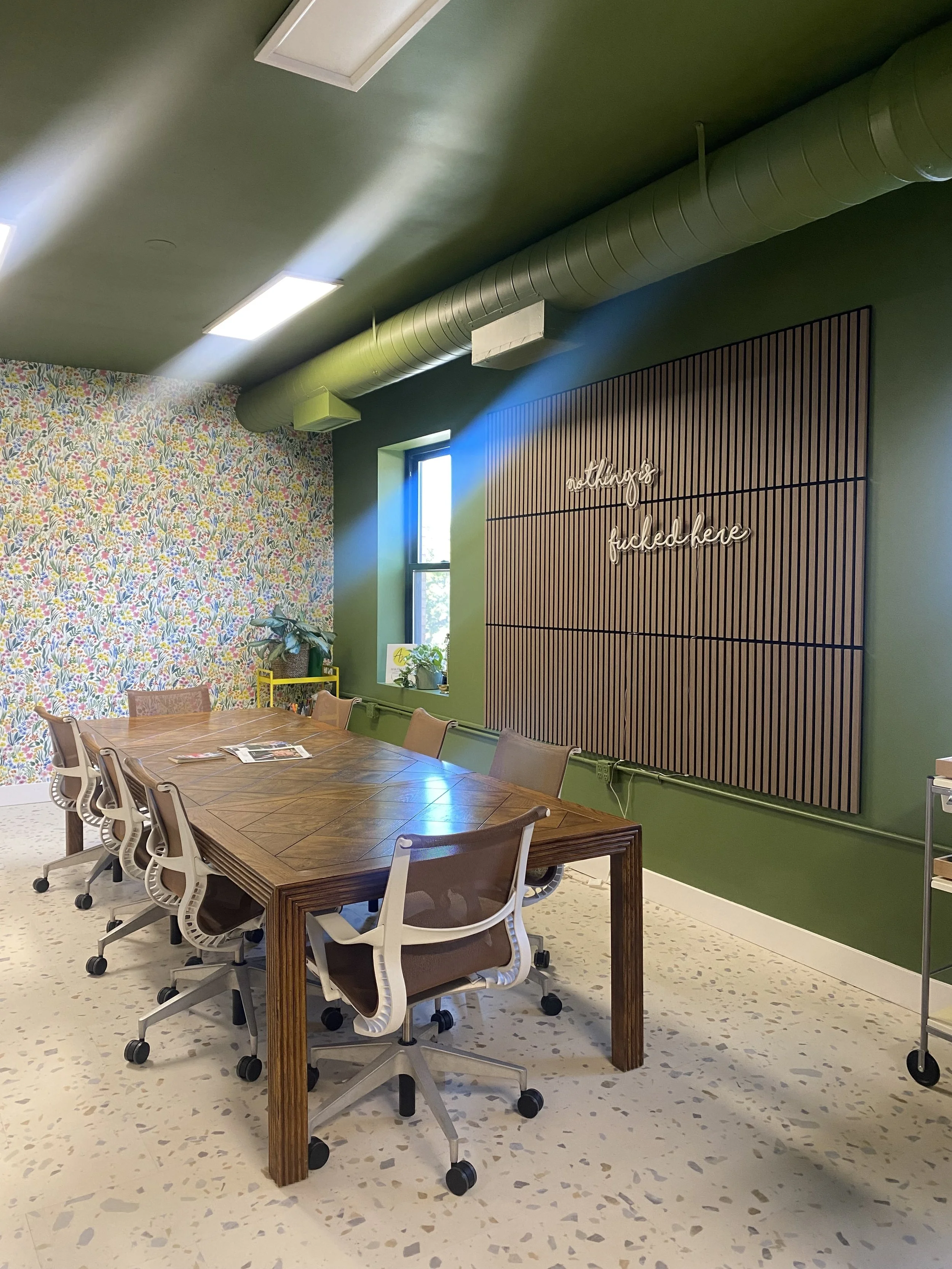 A conference room with a wooden table, eight office chairs with beige cushions, a window with a potted plant on the sill, a floral accent wall, a green wall with a wooden slat decor and neon sign that says 'nothing is fucked here', green ductwork on the ceiling, and speckled white flooring.