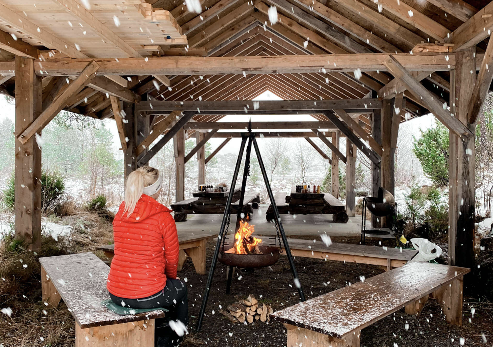 A woman in a red jacket sitting on a wooden bench under a shelter, facing a small fire in a fire pit, with snow falling around and a snowy landscape in the background.