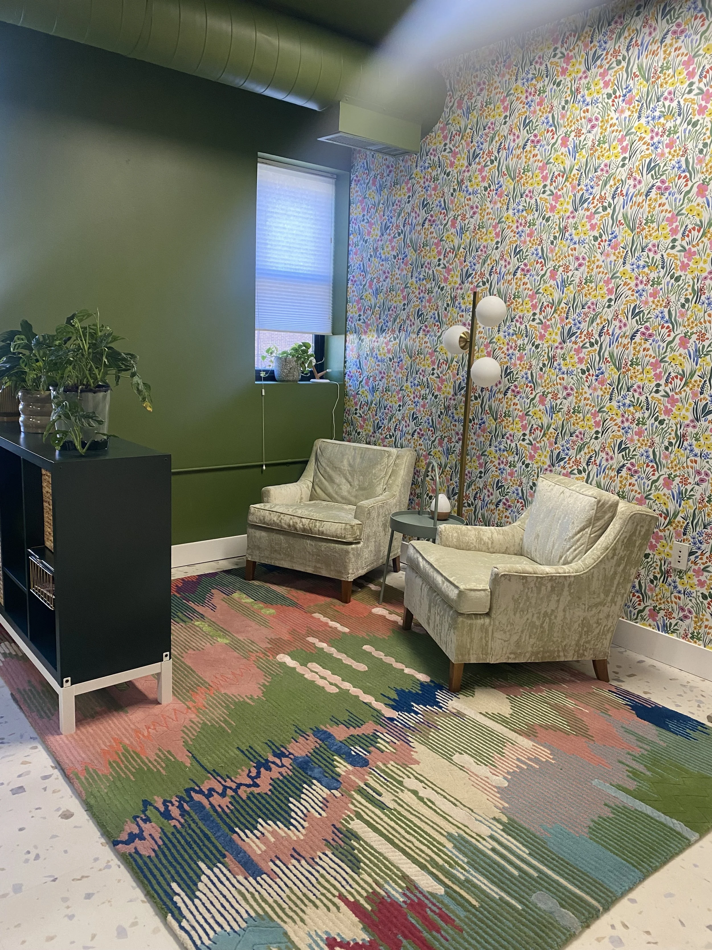 A colorful, cozy room with floral wallpaper, two light green velvet armchairs, a patterned area rug, a standing lamp with multiple bulbs, a small side table, and a window with a blind.
