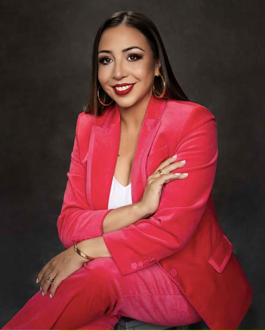 A woman with long dark hair and makeup, smiling with red lipstick, wearing gold hoop earrings, a white top, and a coral pink suit, posing against a dark gray background.