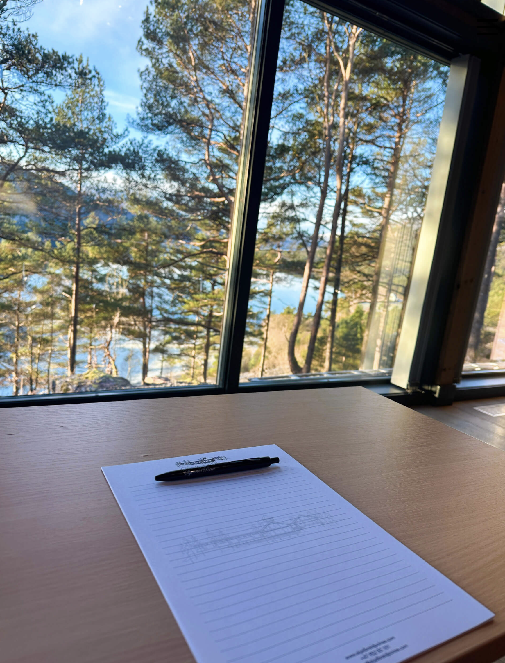 A blank lined notepad with a black pen resting on top is placed on a wooden surface in front of a large window. Outside the window, tall pine trees and a body of water are visible, indicating a forested lakeside setting during daytime.