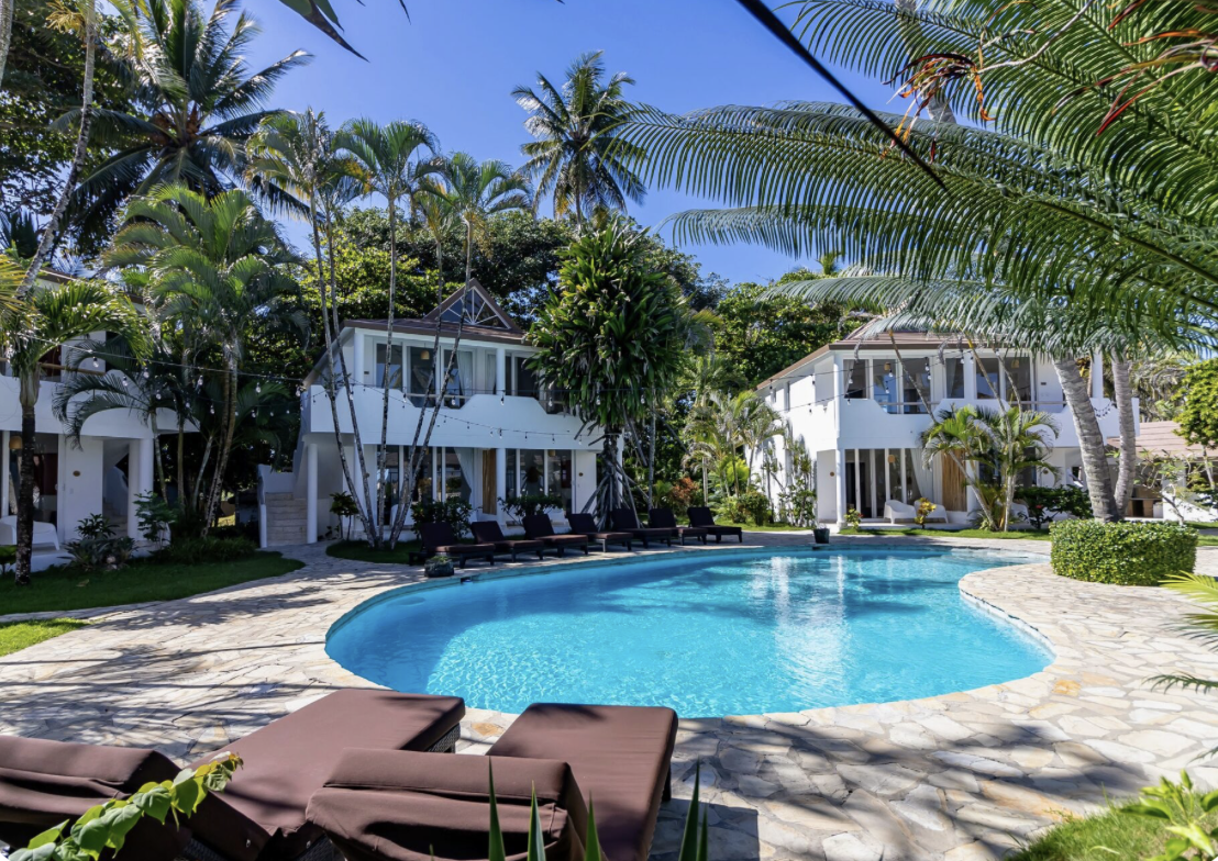 A tropical resort with two white buildings and a large swimming pool surrounded by lounge chairs and lush palm trees under a clear blue sky.