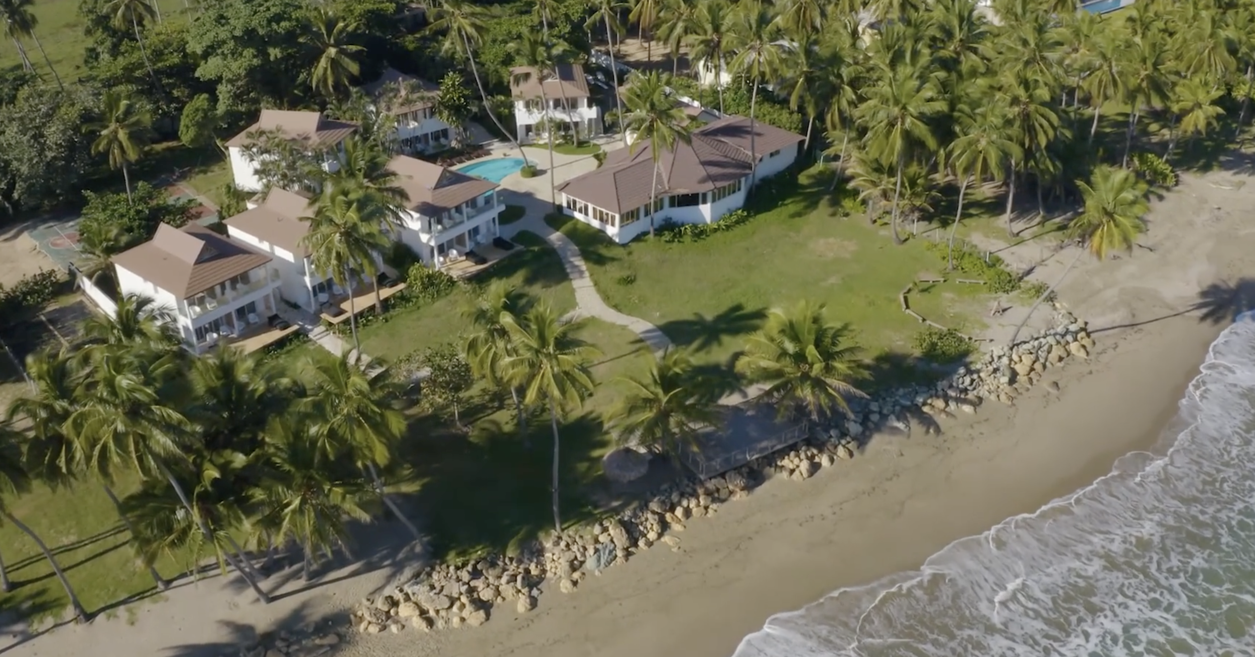 Aerial view of beachfront cottages with palm trees and a swimming pool, beach and ocean waves in the background.