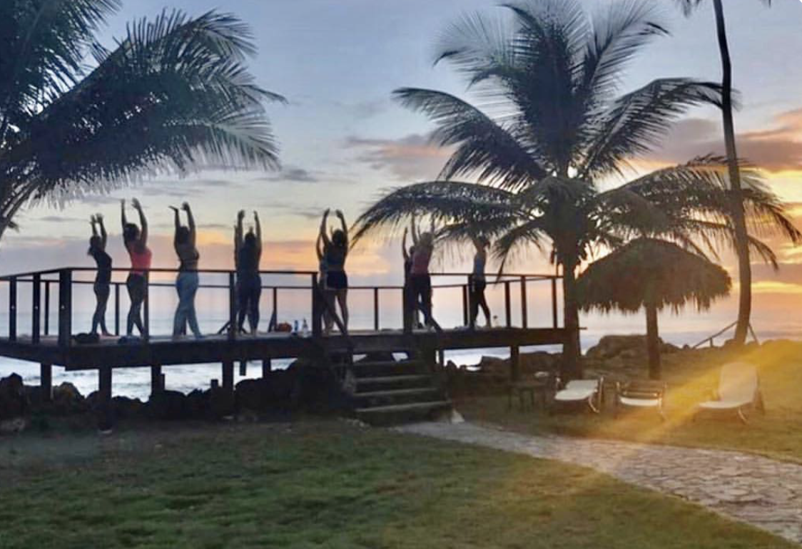 Group of people practicing yoga on a wooden deck near the beach at sunset, with palm trees and ocean in the background.