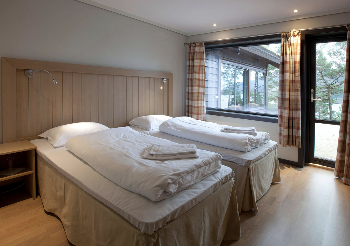 A bedroom with two beds, each with white bedding and towels, a wooden headboard, a nightstand, and a large window with curtains showing trees outside.