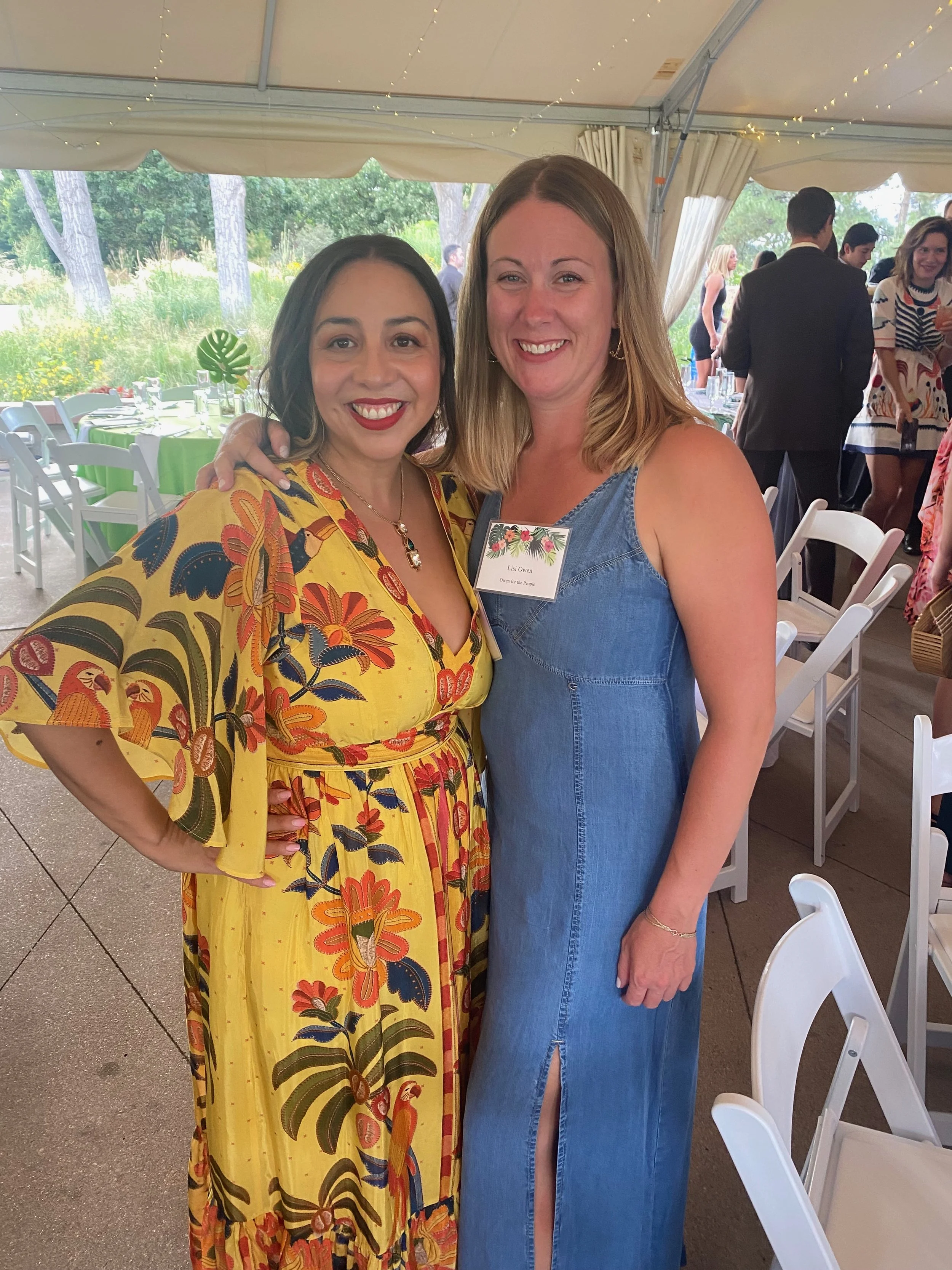 Two women smiling at a social gathering under a tent, with decorated tables and other people in the background.
