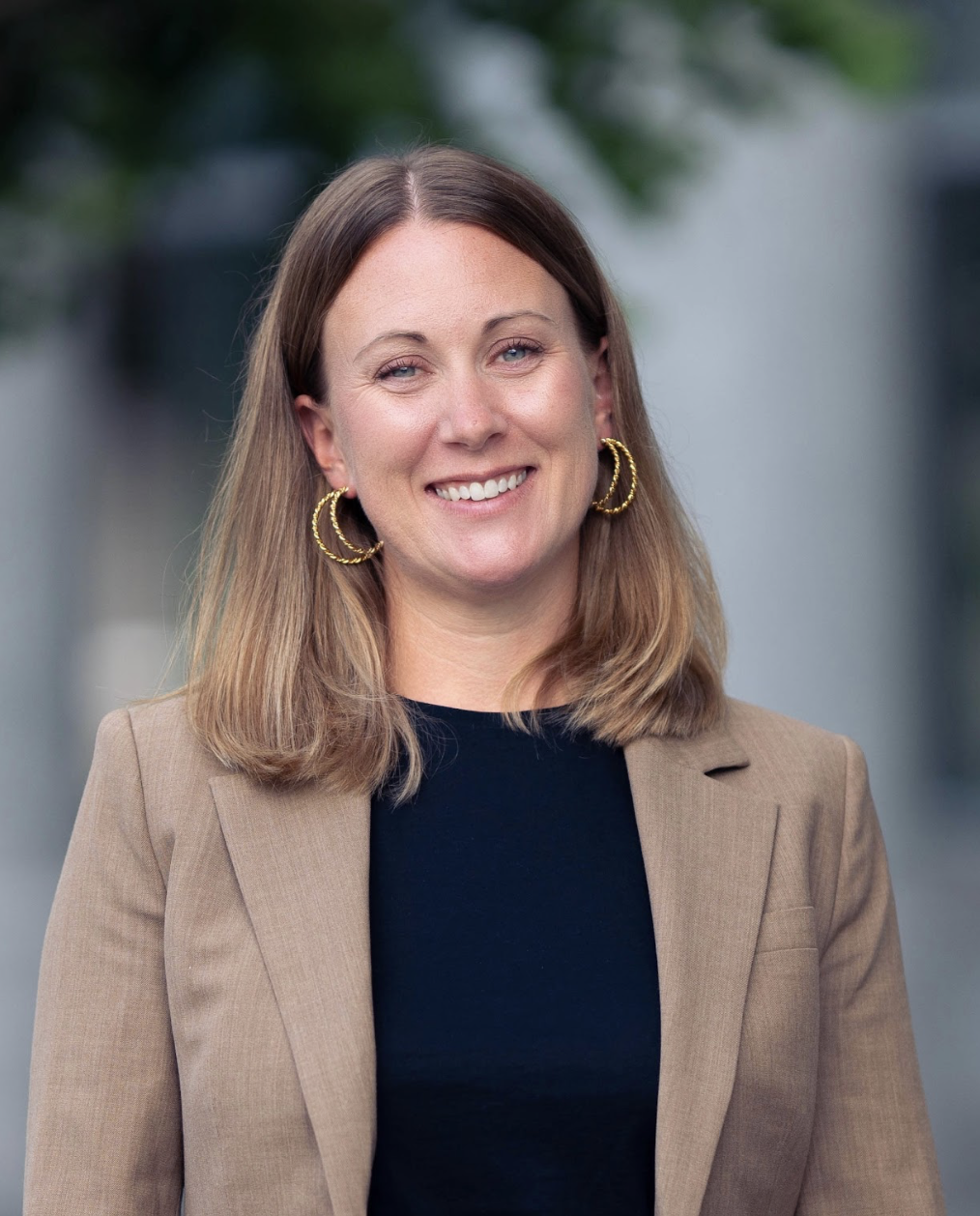 A woman with shoulder-length light brown hair, wearing a tan blazer and black top, smiling outdoors with blurred background and green foliage.
