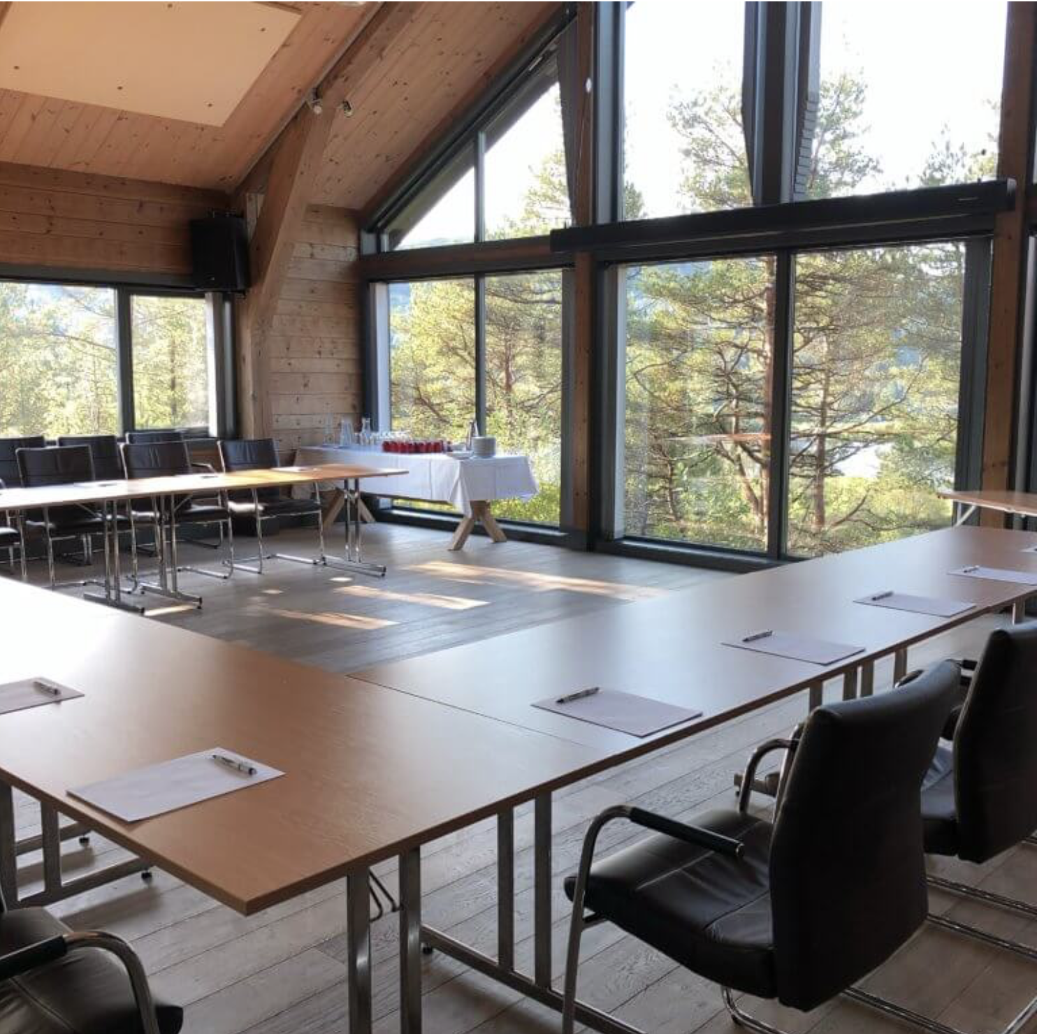 A conference room with tables arranged in a U-shape, black leather chairs, and a small table with glasses and water bottles near large floor-to-ceiling windows showing trees outside.