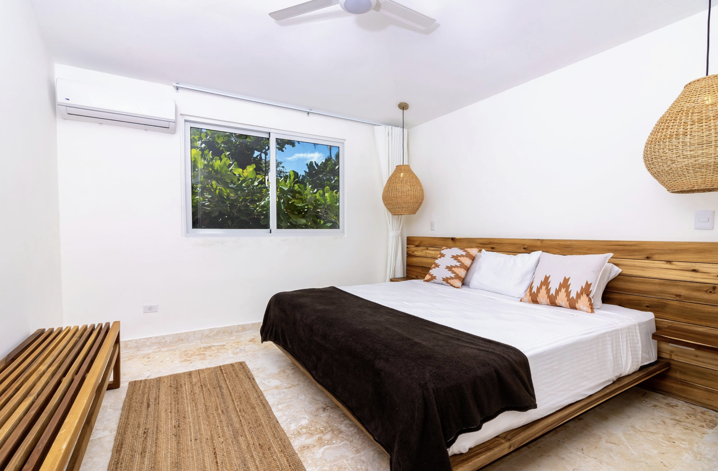 A bedroom with a large bed featuring white sheets, brown and white pillows, and a dark brown blanket, a wooden headboard, wicker pendant lights, a window with greenery outside, and a wooden bench on a beige marble floor.