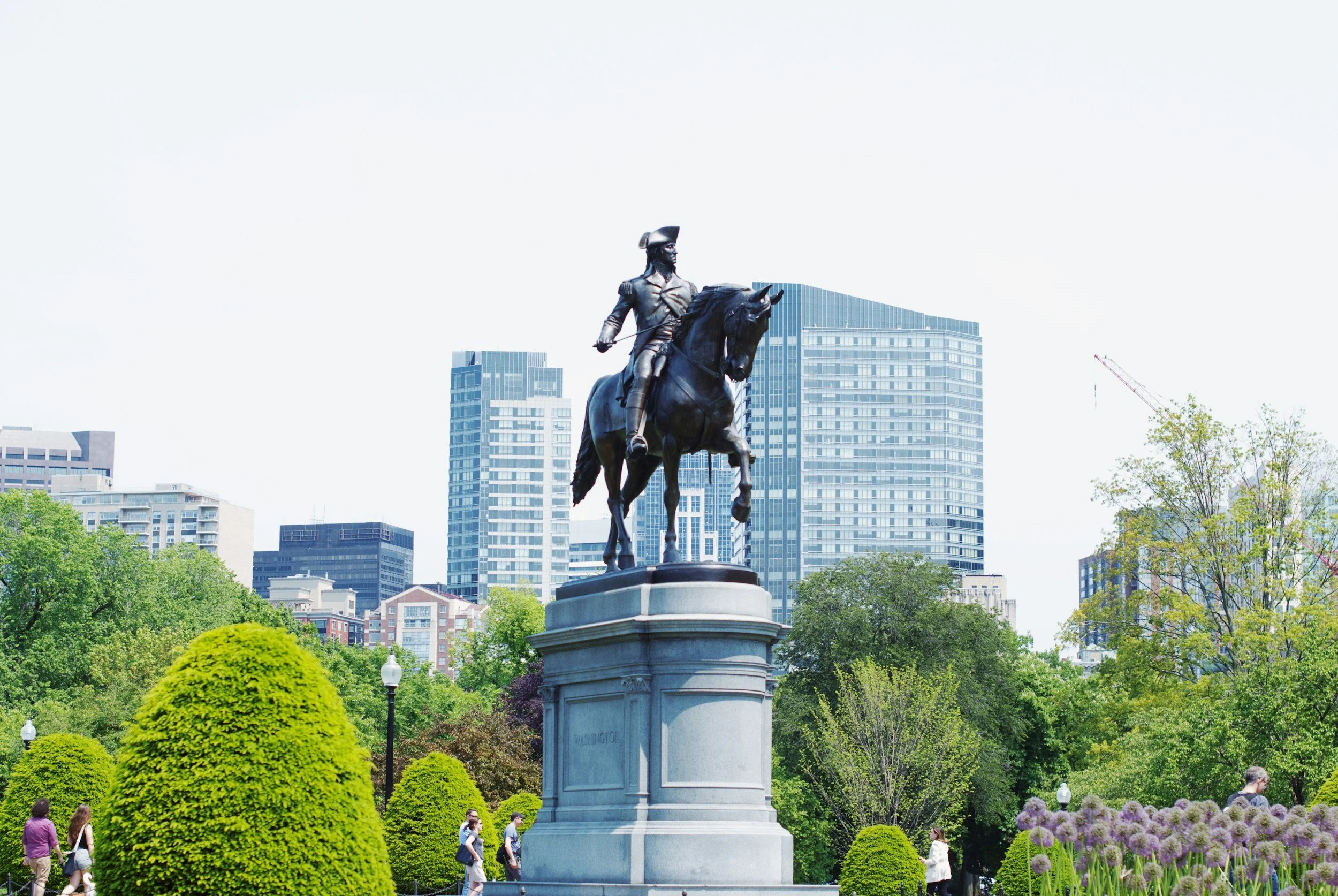 Equestrian statue of a historical figure on horseback in a city park, with tall buildings and green trees in the background.
