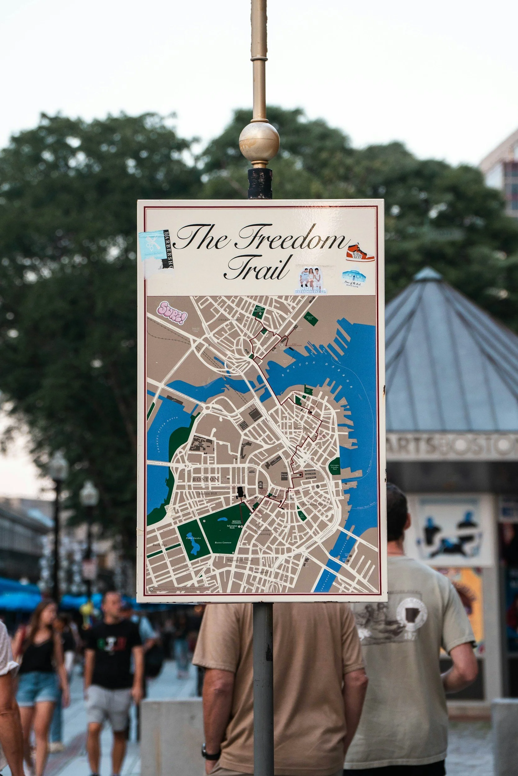 City map signage titled 'The Freedom Trail' with various landmarks and paths, surrounded by pedestrians in an outdoor area.