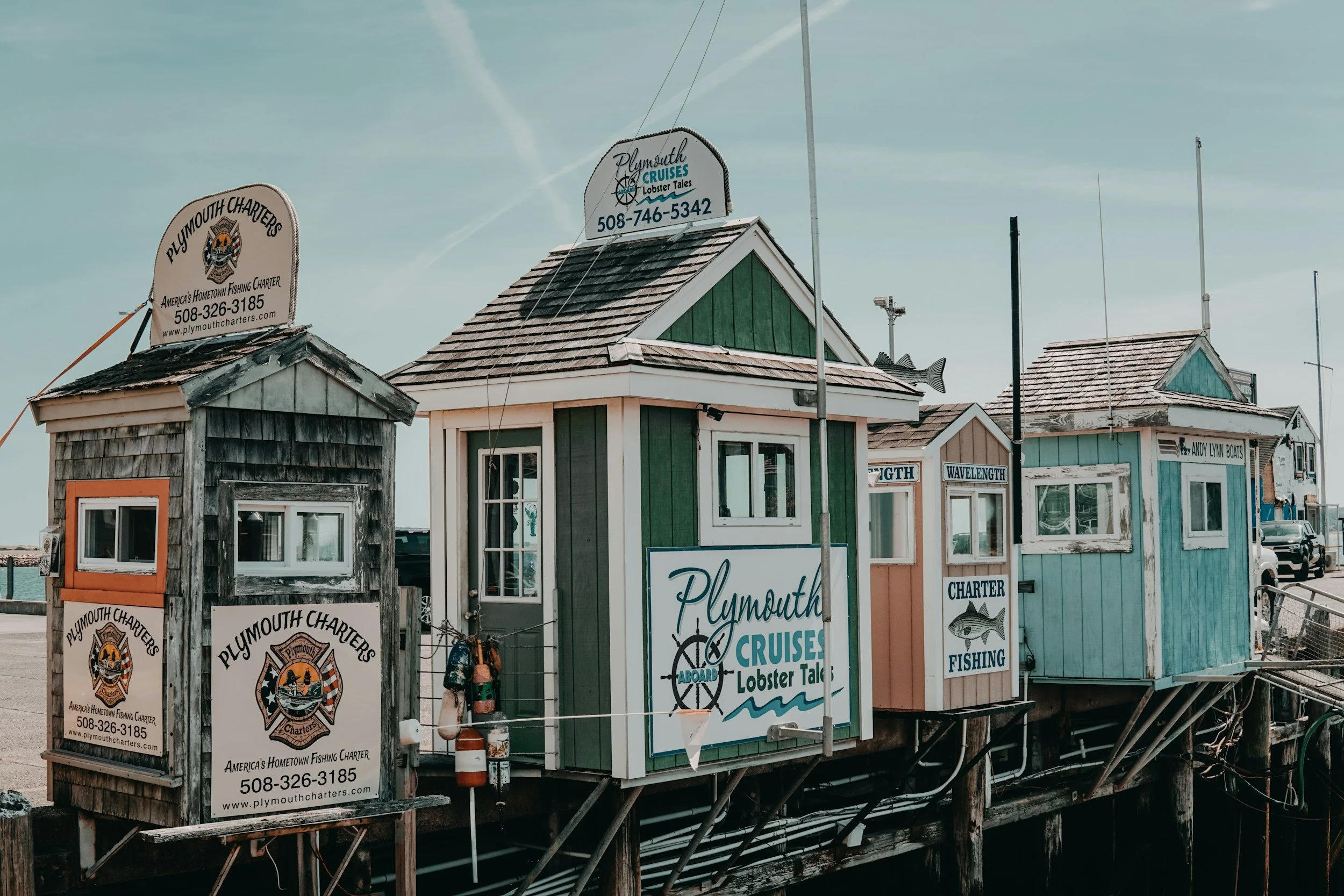 Colorful small wooden buildings on stilts at a dock, with signs advertising Plymouth Charters, cruises, lobster tales, and fishing, under a partly cloudy sky.