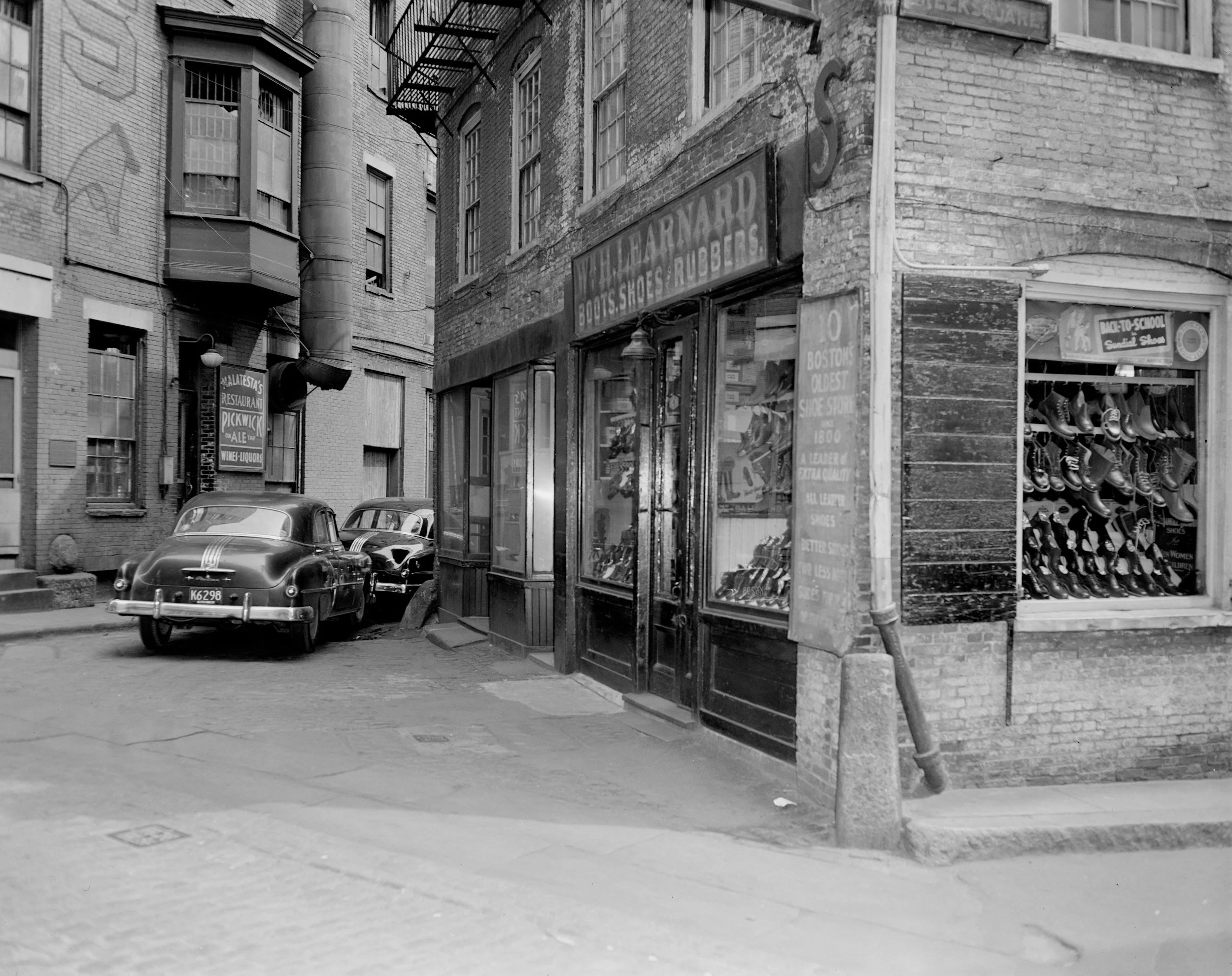 Black and white image of a vintage corner street scene with old cars parked beside a shoe store and a restaurant in a city block.
