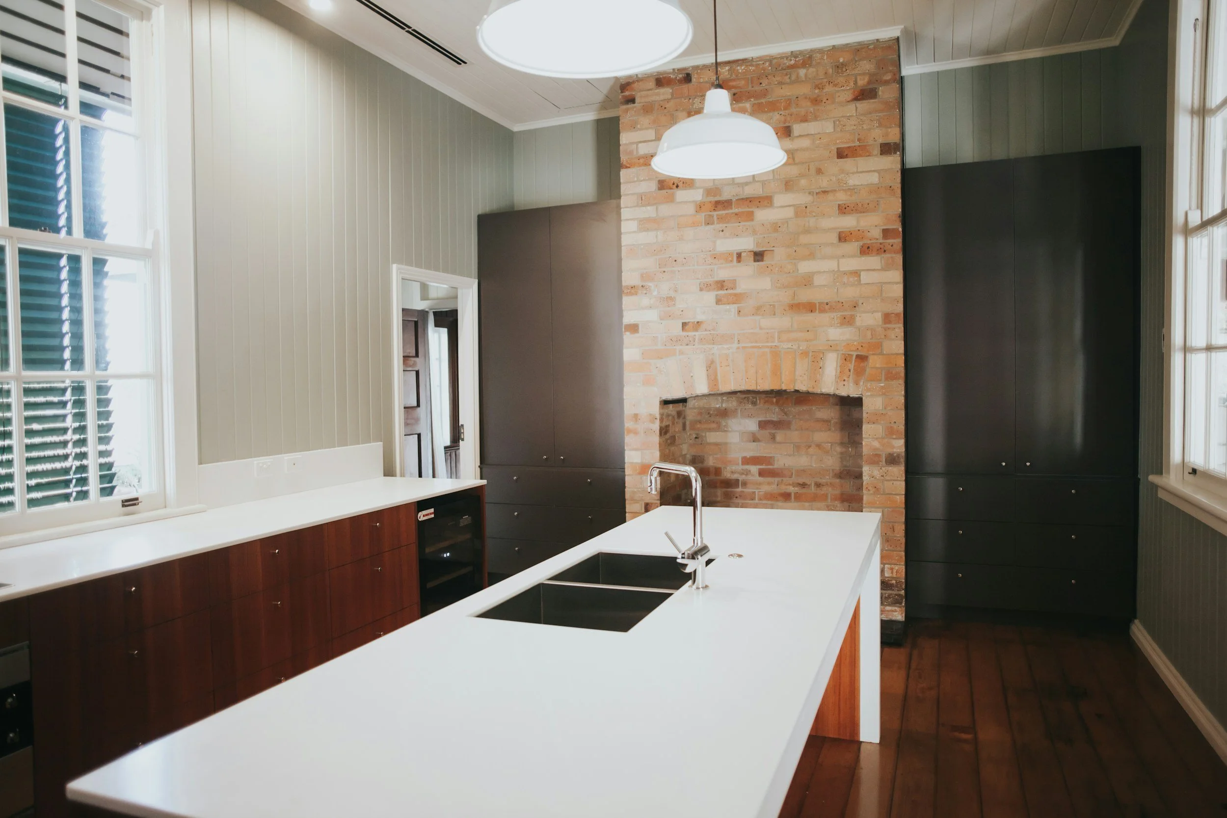 Modern kitchen with white countertops, black cabinets, brick fireplace, wooden floor, and large windows with shutters.