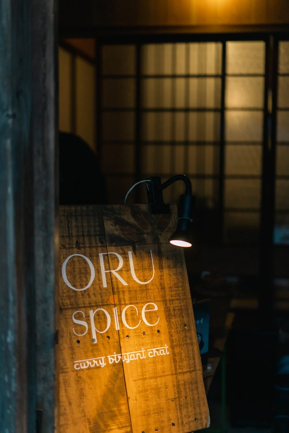 木製の看板に「ORU spice」と書かれていて、下に「curry tiryanti chat」と記載されている。背景は暗い照明の店内で、上部に格子模様の障子のような壁紙が見える。