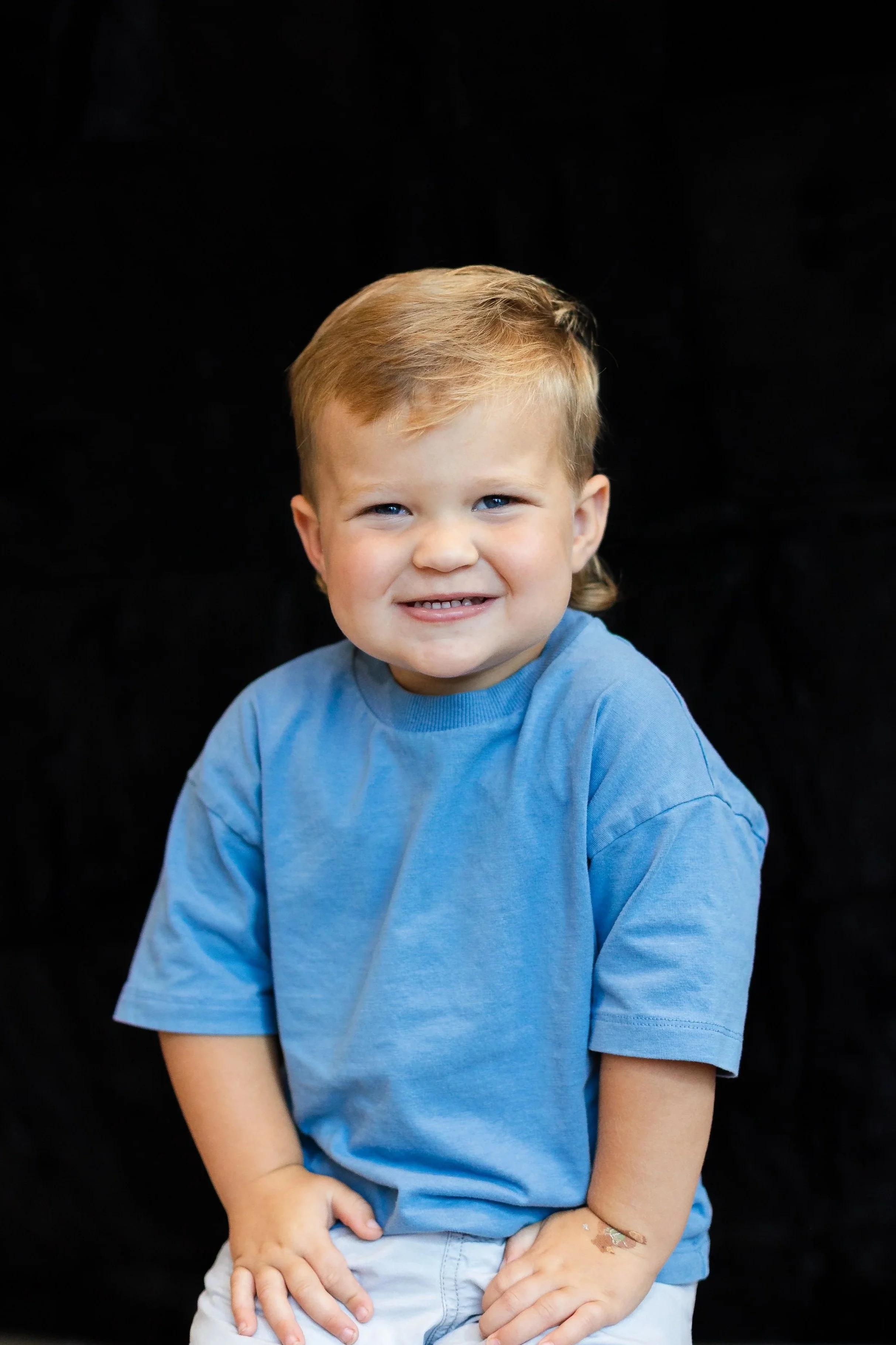 A young boy with blond hair, smiling, wearing a blue t-shirt, against a black background.