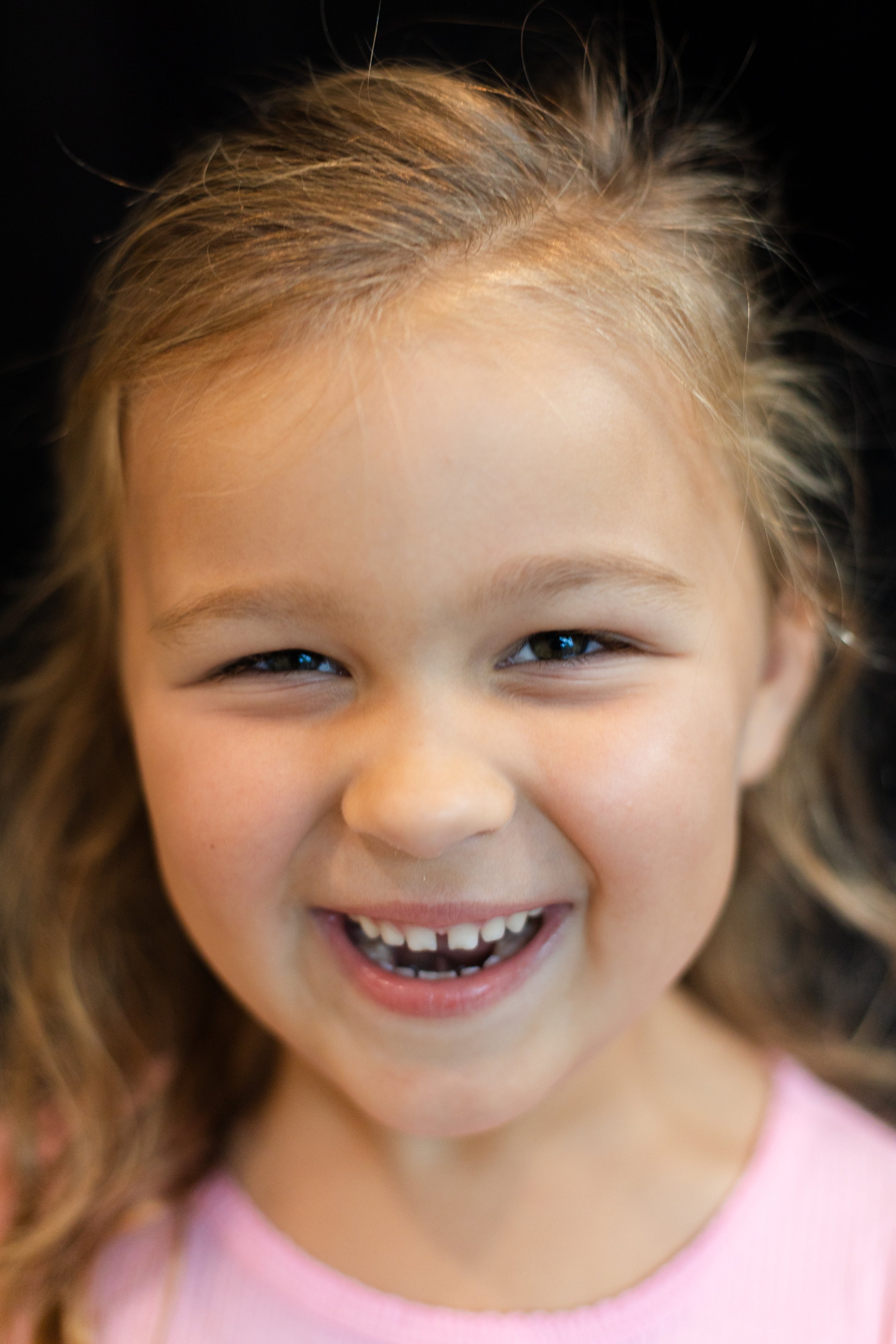 Close-up of a young girl smiling with visible teeth, wearing a pink top, with light brown hair and a dark background.