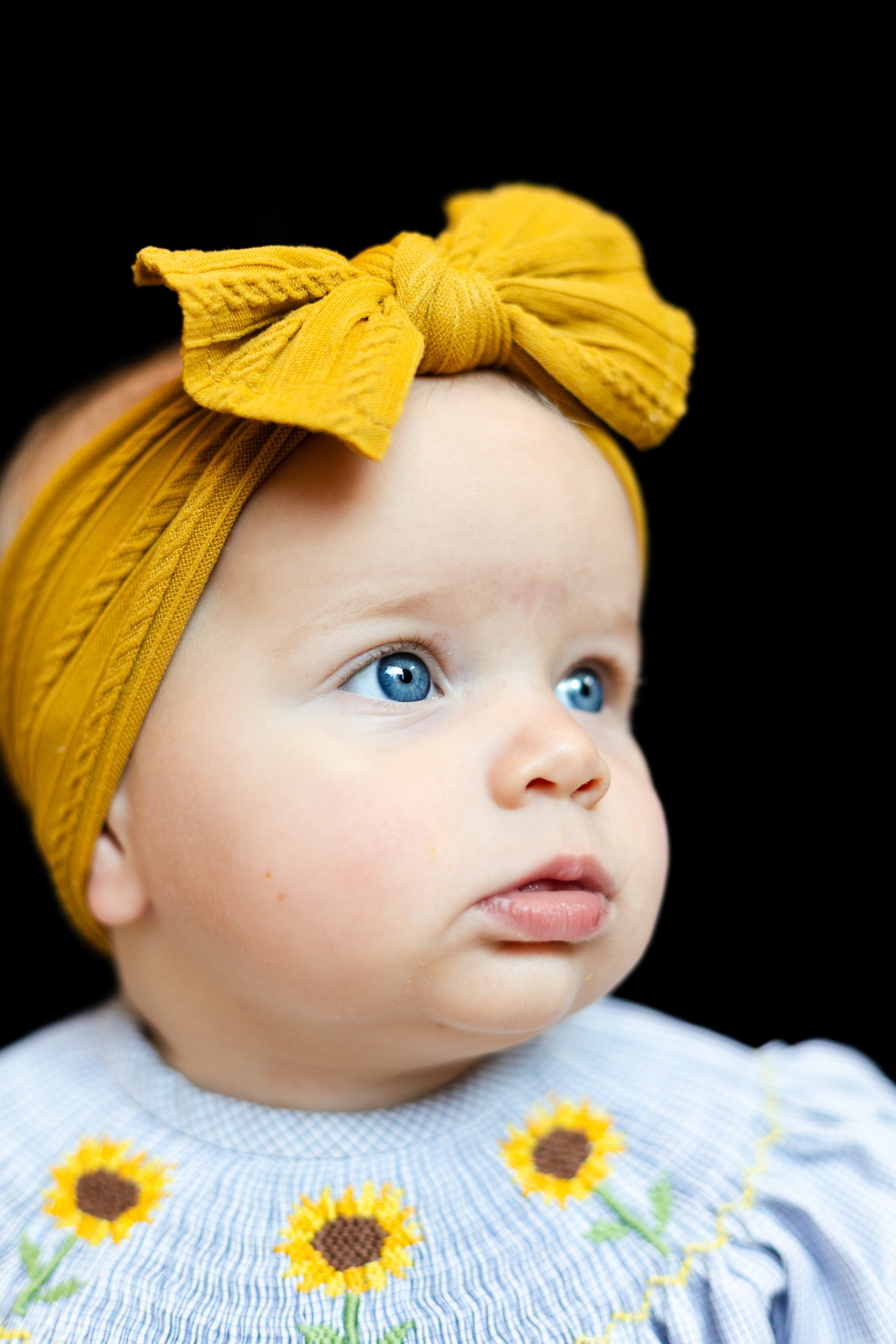 A close-up of a young child with blue eyes, wearing a yellow headband with a bow, and a dress with sunflower embroidery.