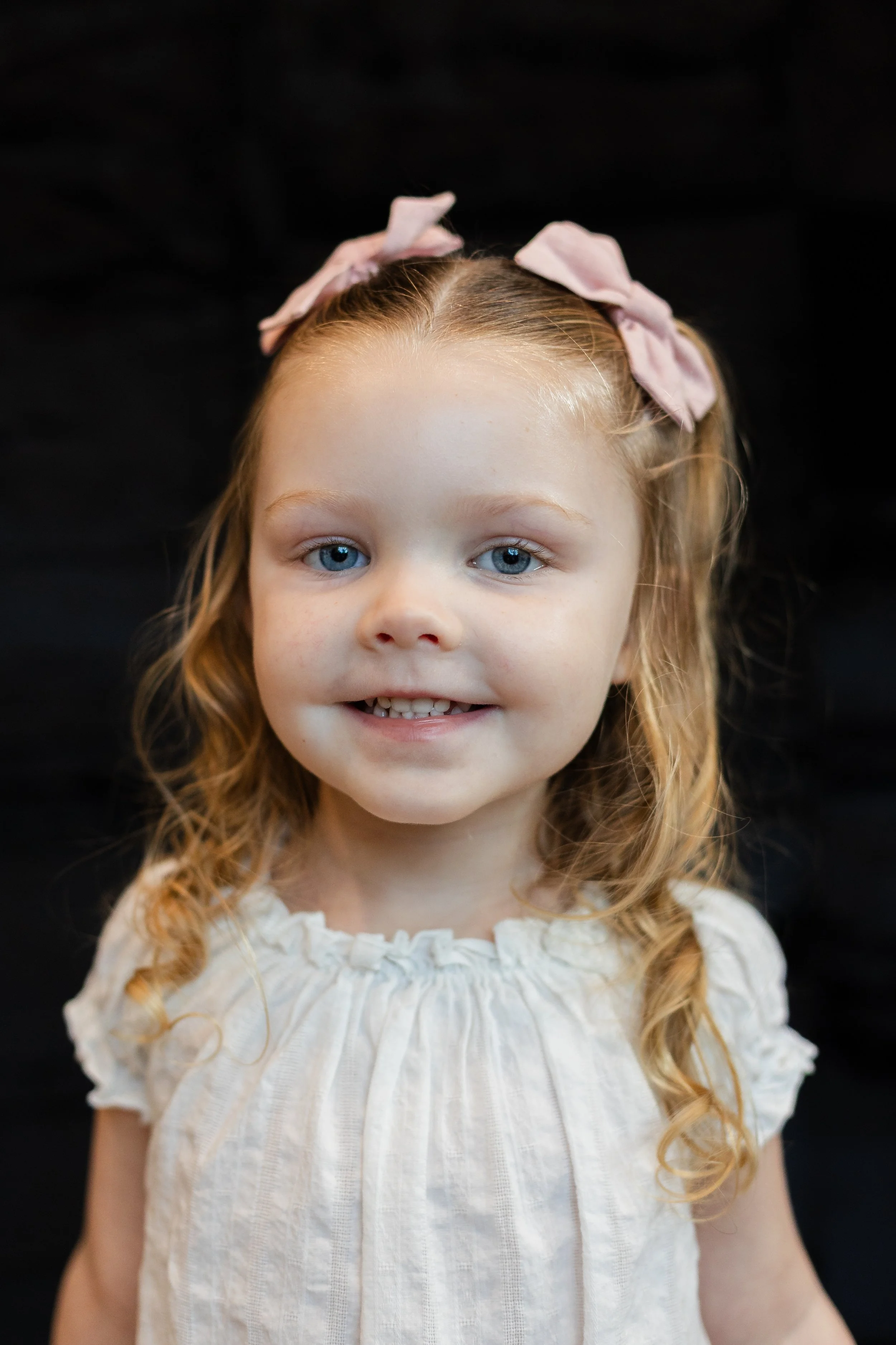 A young girl with red hair and blue eyes smiling, wearing a white ruffled top and a pink bow headband against a black background.