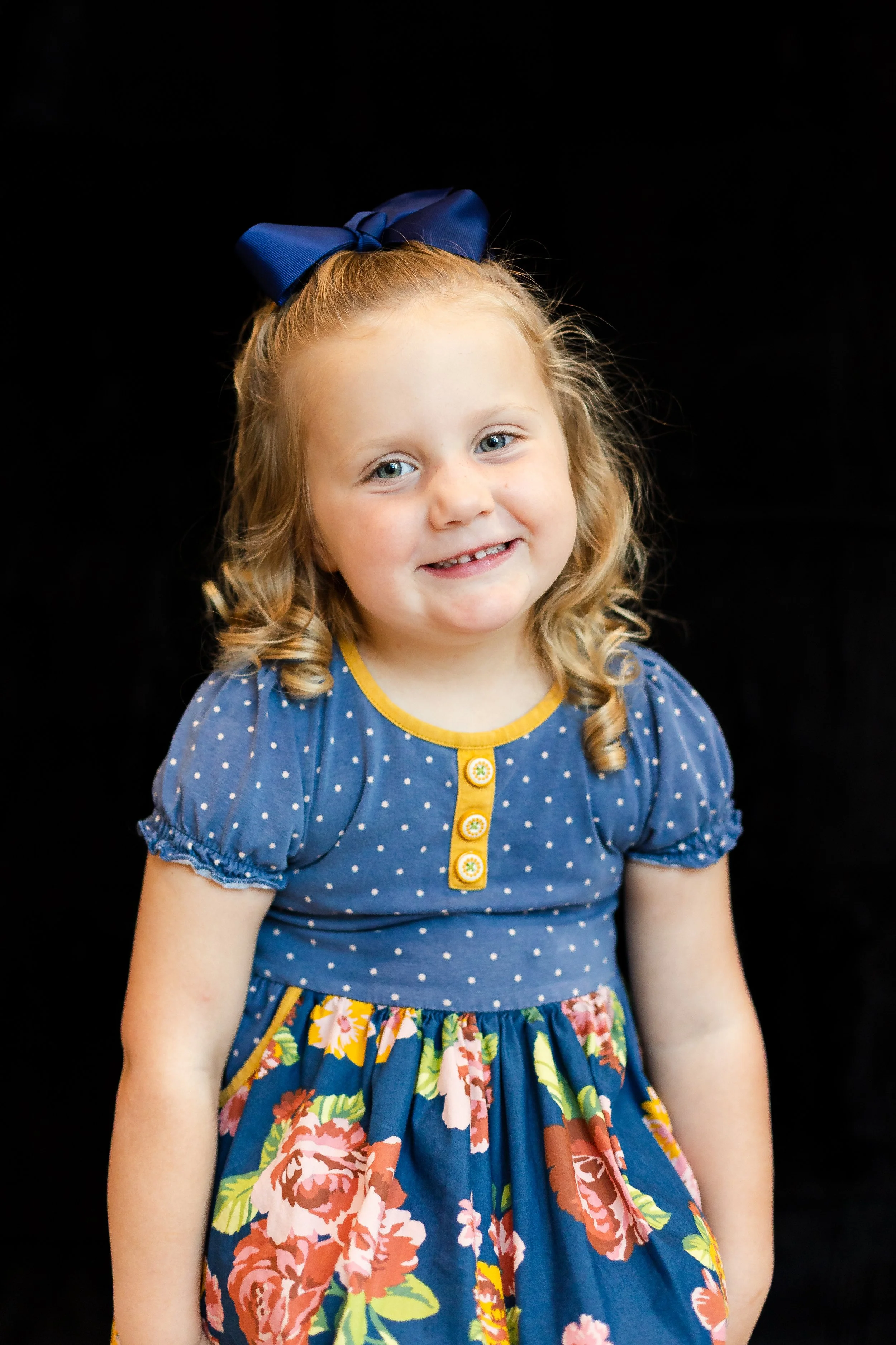 A young girl with blonde curly hair, wearing a blue dress with white polka dots, a yellow trim, and floral patterns, and a large blue bow in her hair, smiling against a black background.