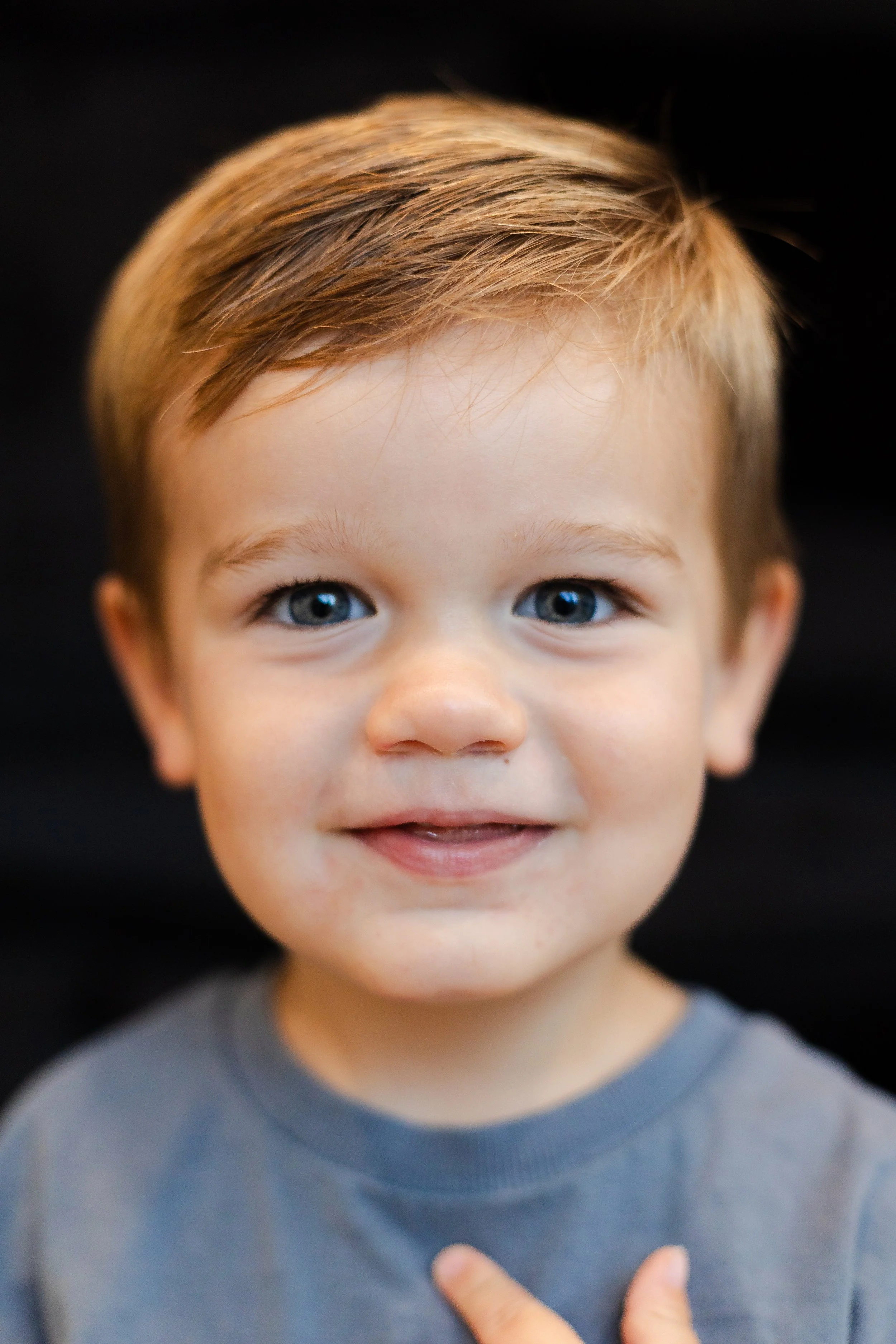 Close-up of a young boy with light brown hair and blue eyes, smiling slightly, wearing a gray shirt, against a black background.